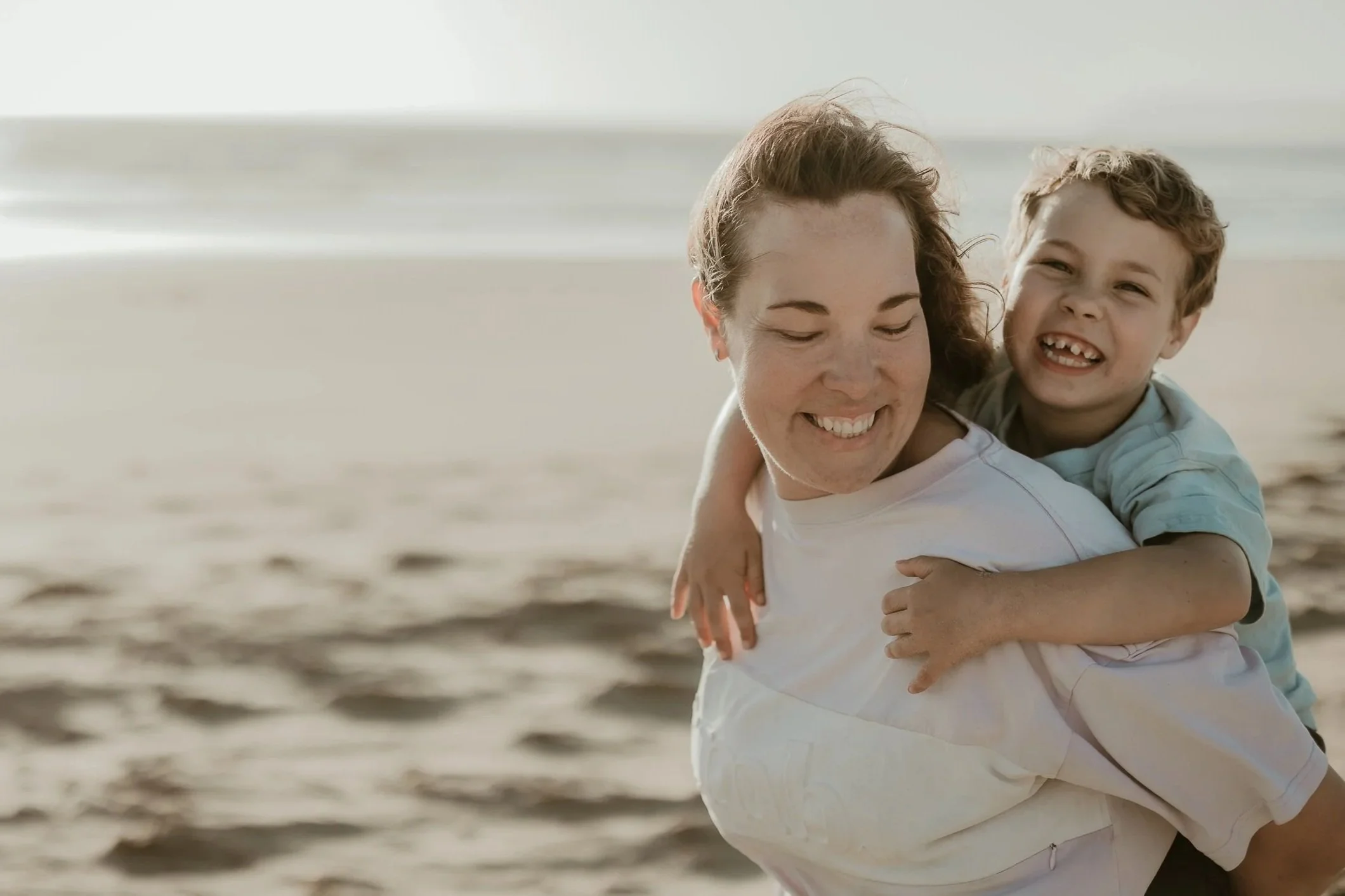 A woman and a young boy smiling and hugging on the beach.