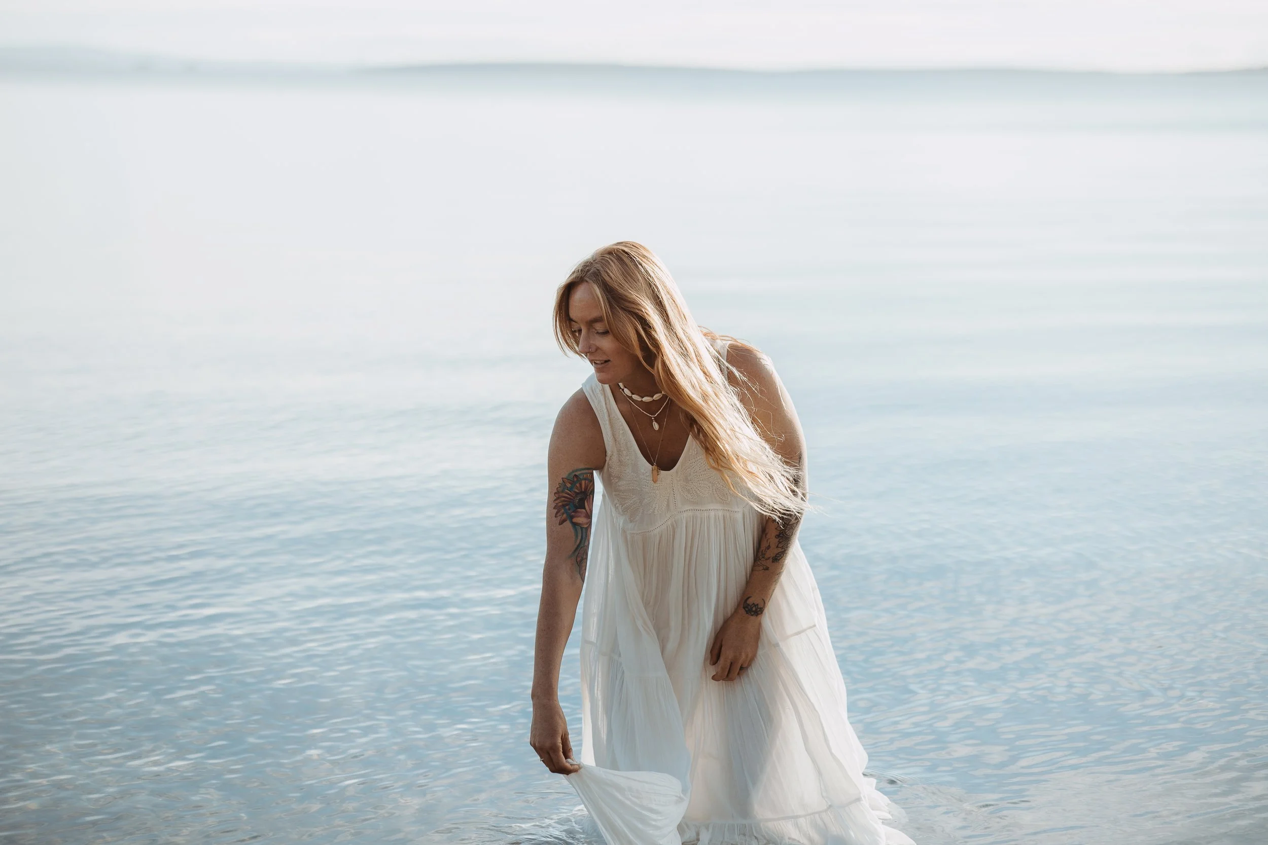A woman standing in shallow water at the beach, wearing a white dress and multiple necklaces, with tattoos on her arms, looking down and holding her dress.