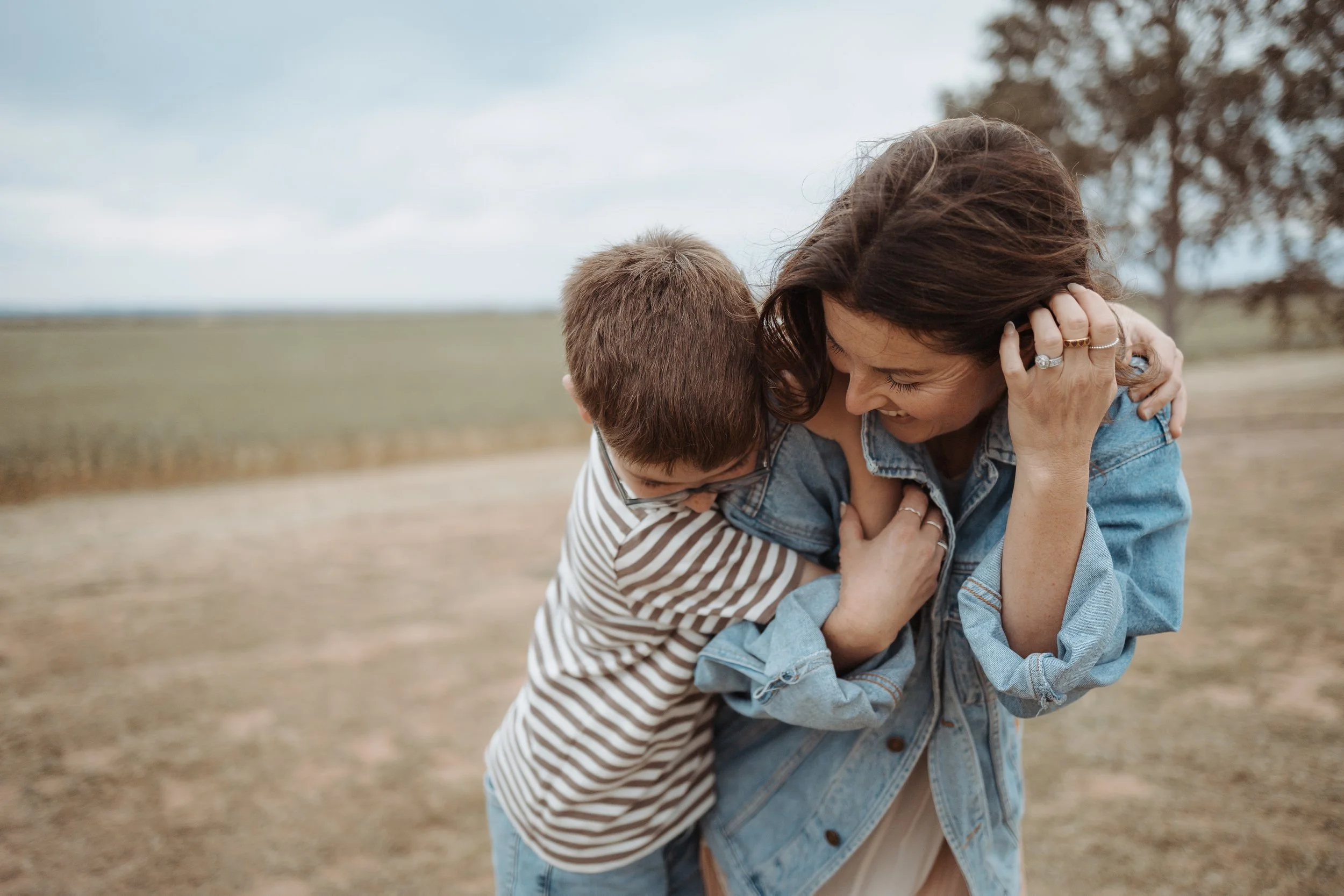 A woman and a young boy hugging outdoors on a cloudy day with an open field and a tree in the background.