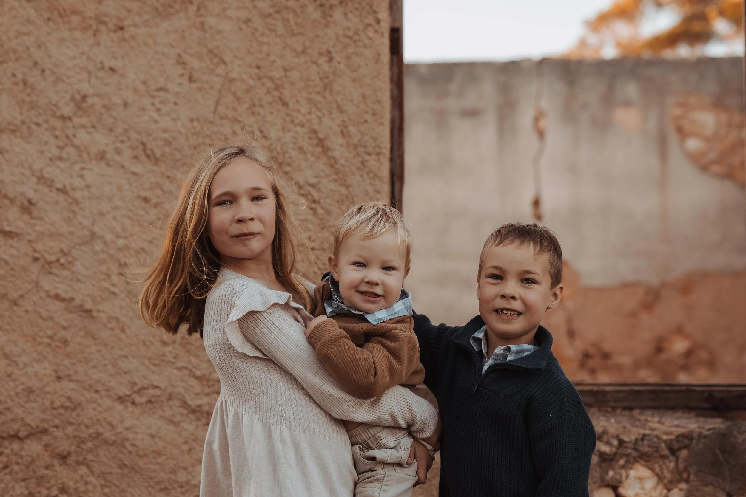 Four children standing outdoors against a textured beige wall, with autumn foliage visible in the background.