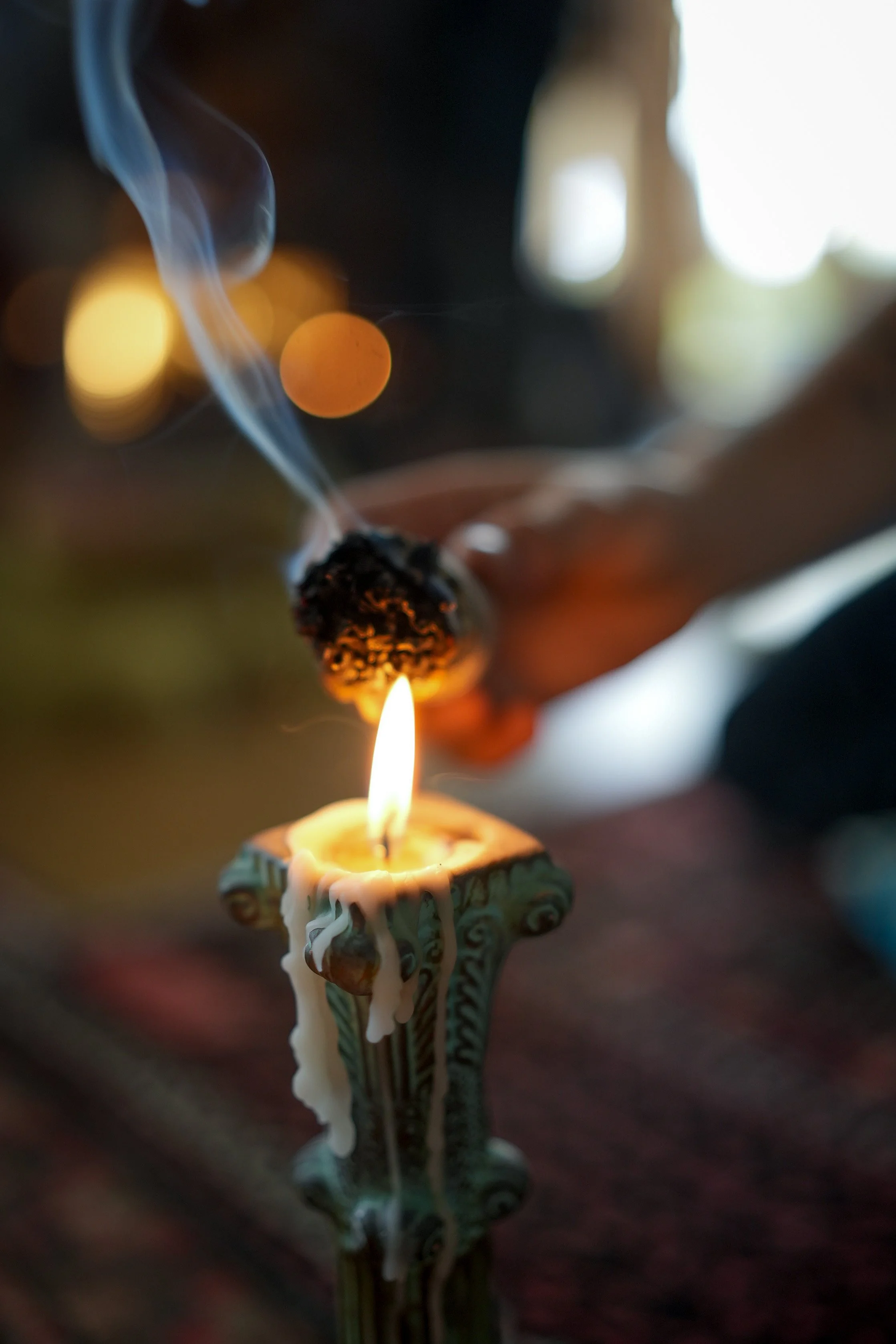 Close-up of a lit candle with melting wax, smoke rising, and a person holding a burning stick with a charred end in the background.