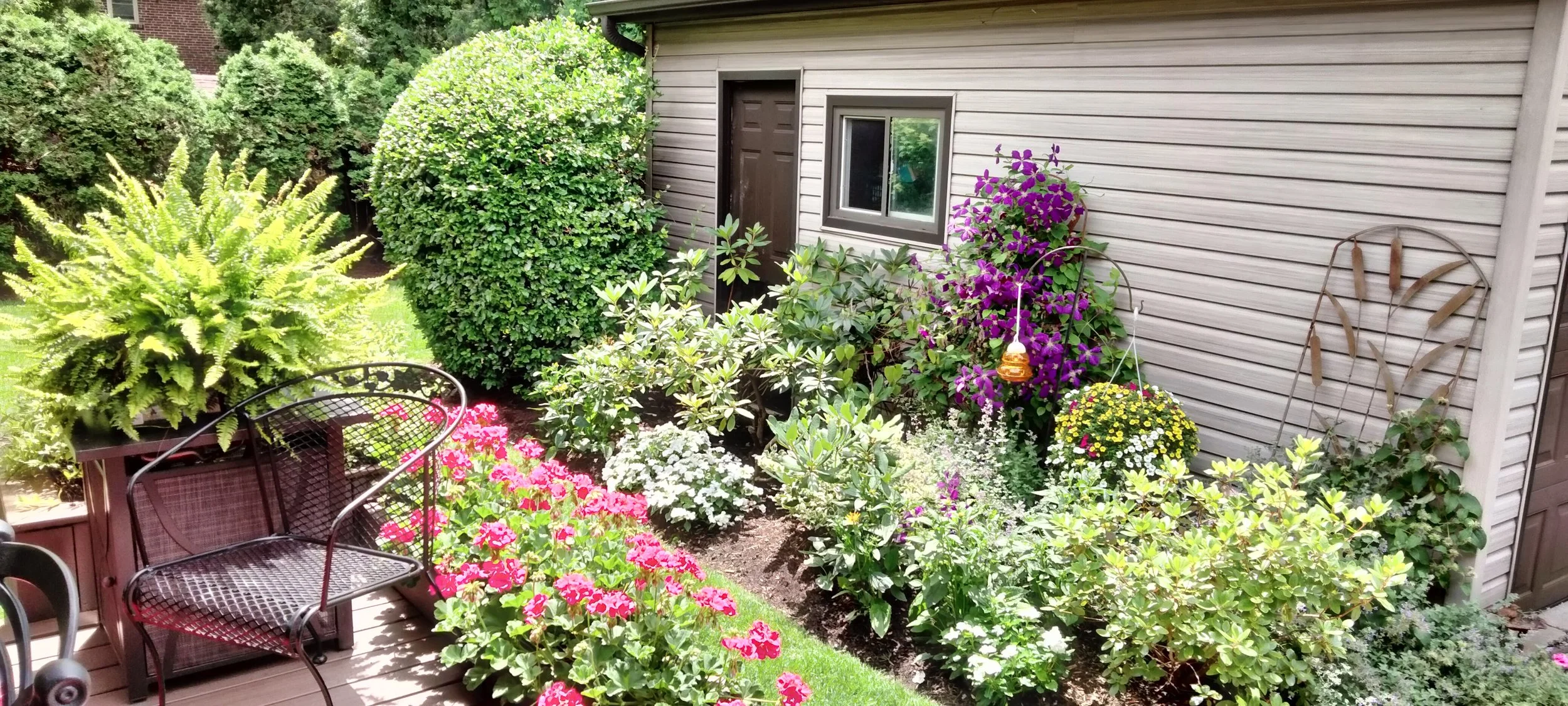 A backyard garden with a variety of colorful flowers, green bushes, and a decorative metal garden ornament against the side of a house with beige siding, a small window, and a brown door.