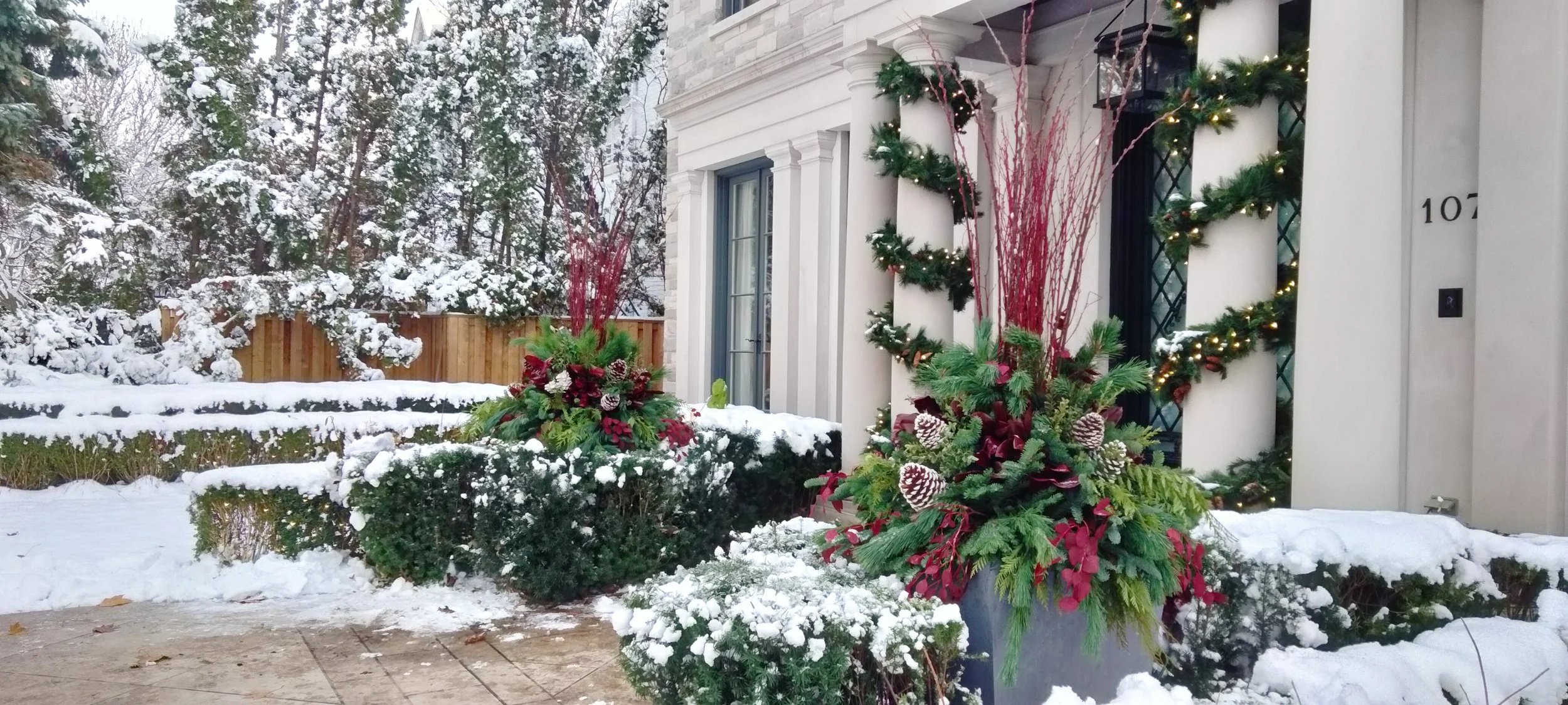 Decorated house exterior with Christmas wreaths and garlands, and holiday floral arrangements, amid snowy landscape.