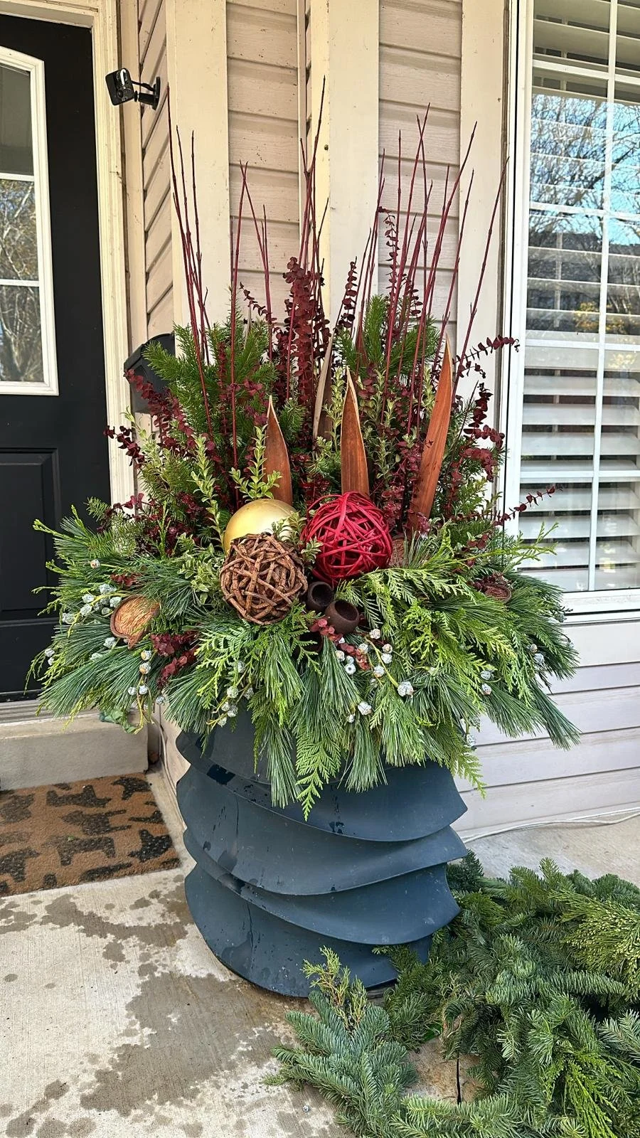 Large holiday floral arrangement with greenery, red and gold ornaments, and decorative twigs, placed outside a house near the front door.