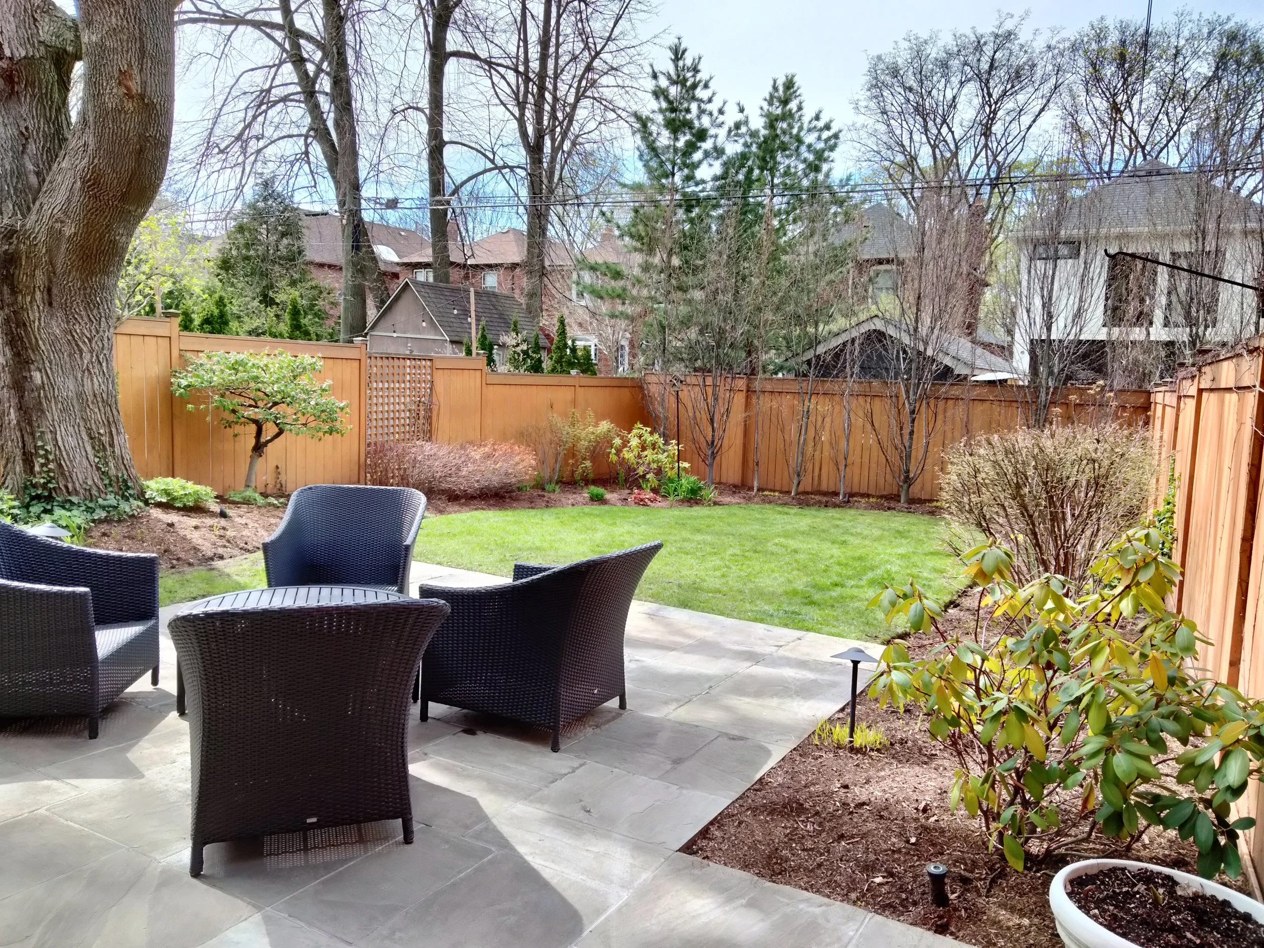View of a backyard with a patio and outdoor furniture, a lawn, trees, shrubs, and a wooden privacy fence.