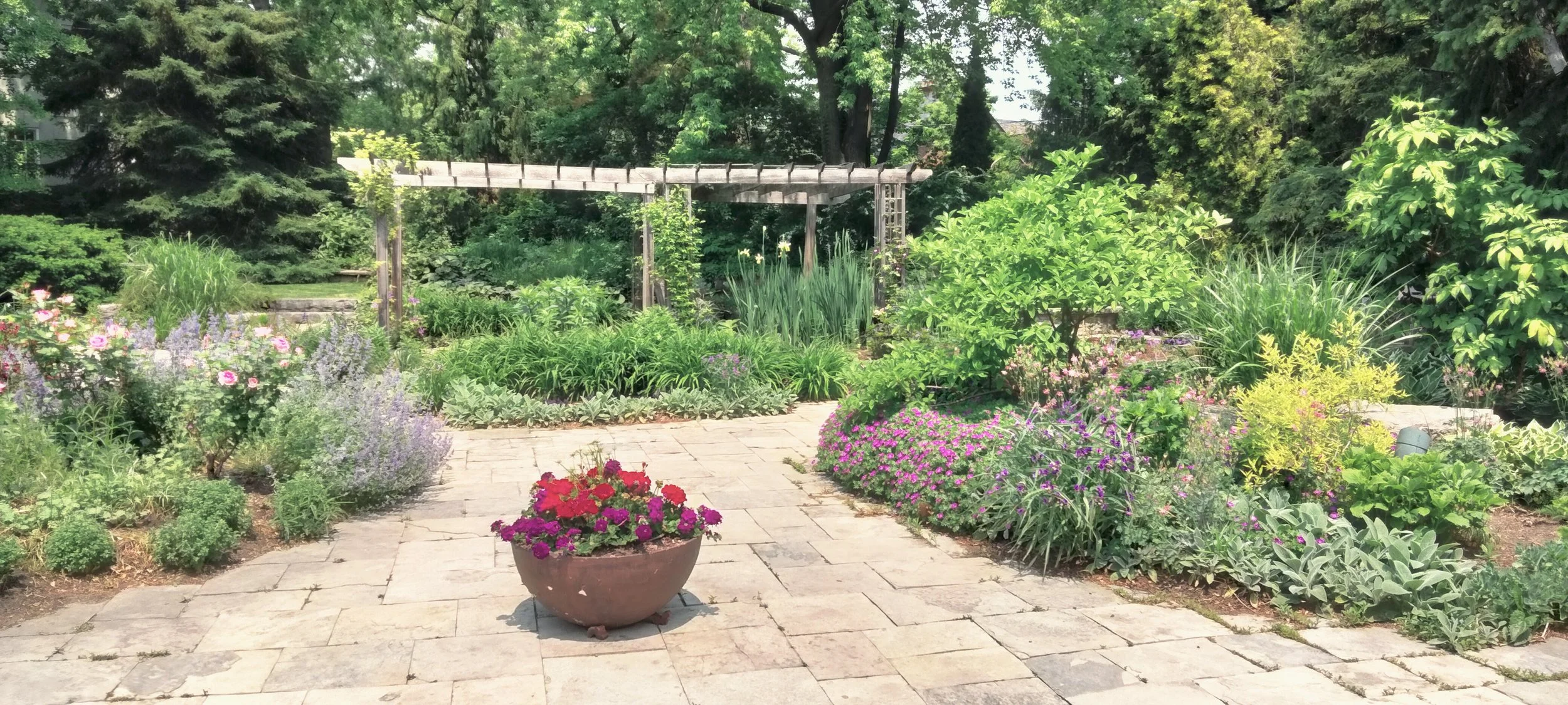 A lush garden with a stone patio, colorful flowers, and green foliage, featuring a small potted plant with red and purple flowers in the foreground, and a wooden trellis in the background surrounded by trees and plants.