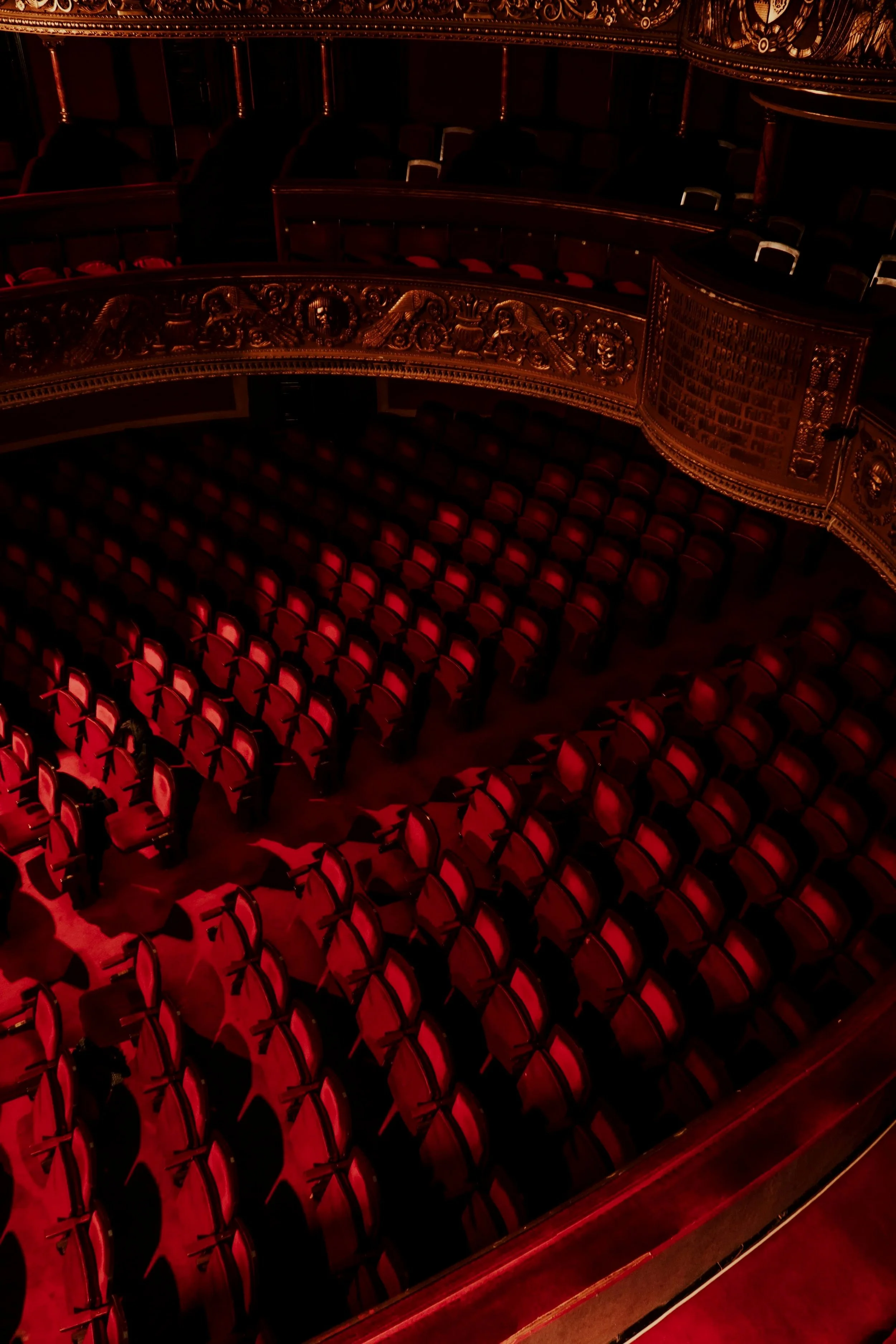 Empty theater with red lights illuminating upholstered seats and ornate gold detailing on the balcony railing.