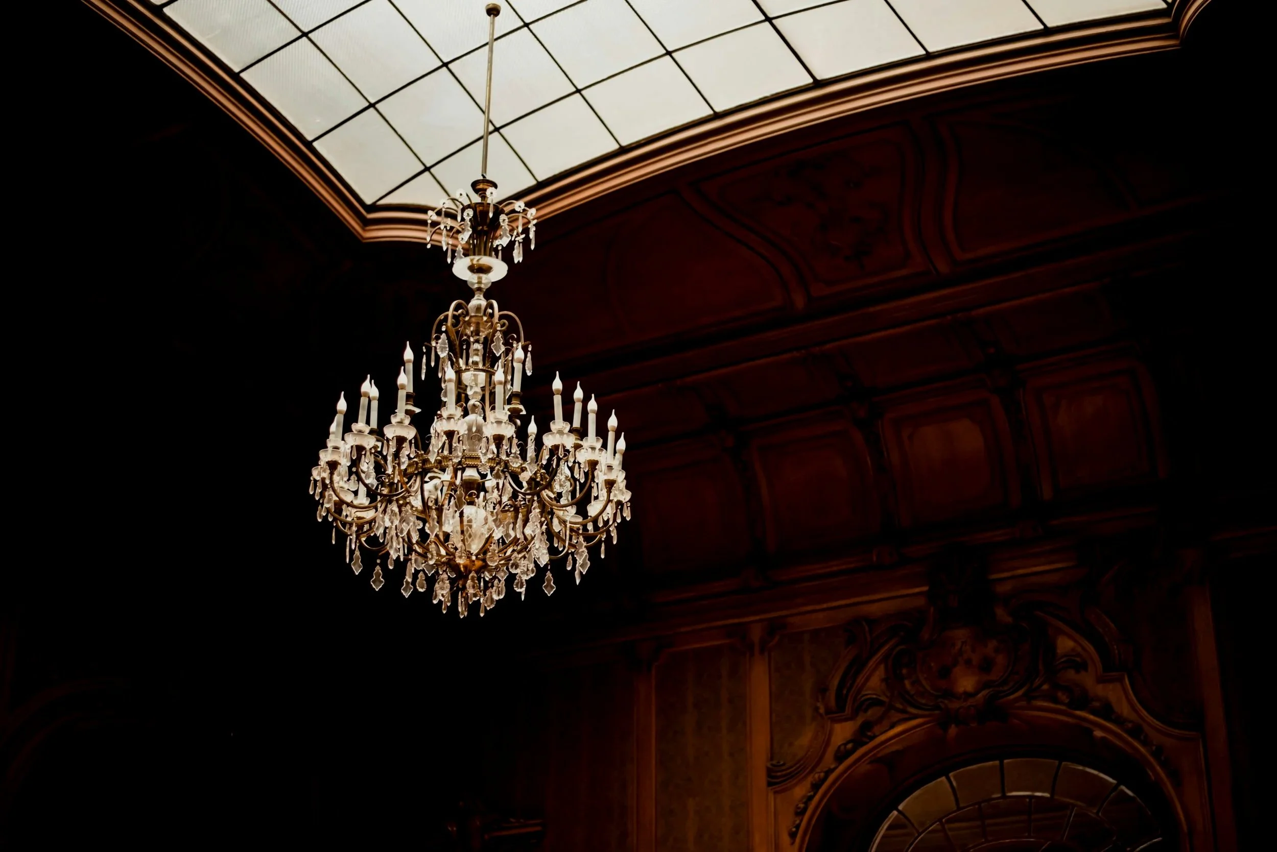 Elegant crystal chandelier hanging from a ceiling with a skylight above, in a richly decorated room with dark wood paneling and a large mirror.