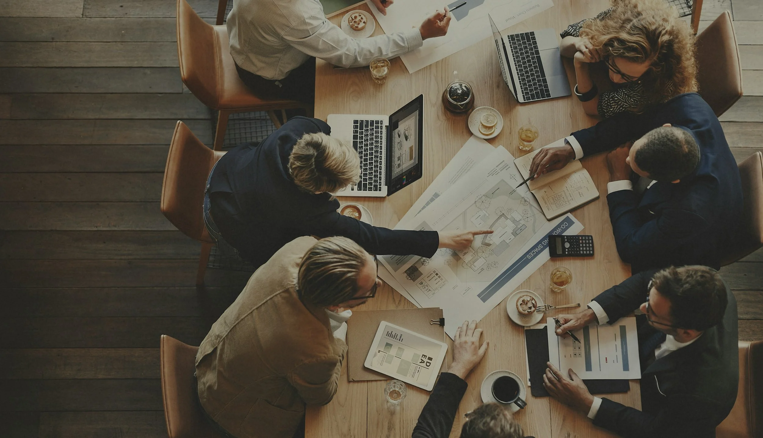 A group of six professionals sitting around a wooden conference table discussing architectural plans, with laptops, documents, and drinks on the table.