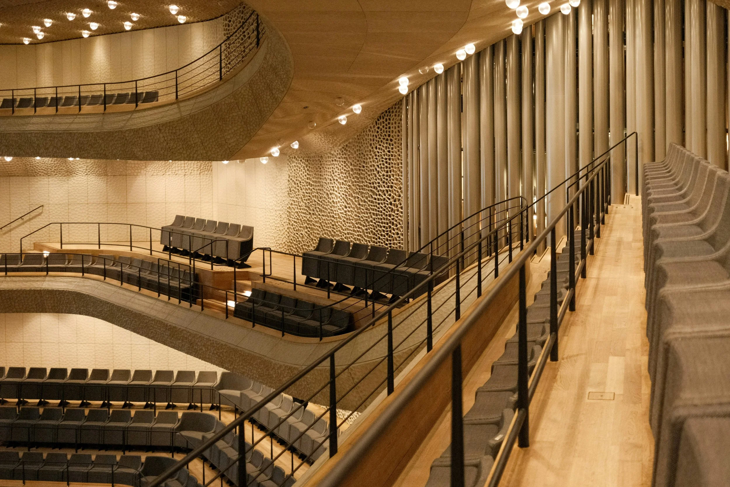 Interior of a modern auditorium or theater with tiered seating, black chairs, wooden flooring, decorative wall panels, and ceiling lights.