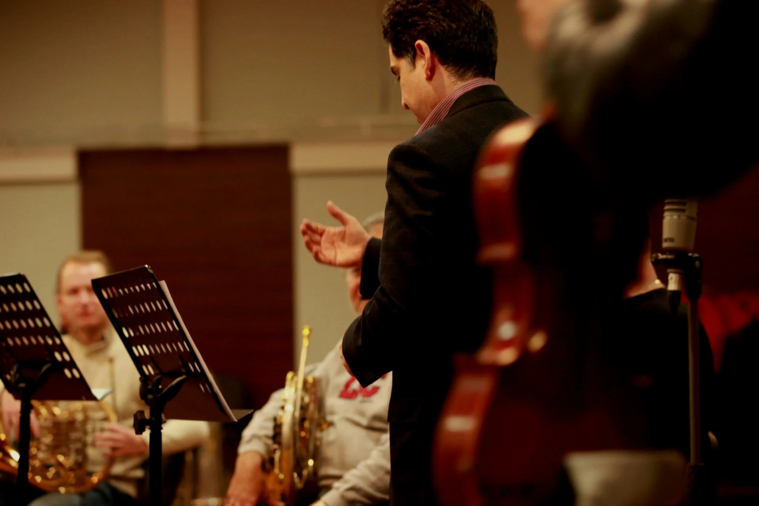 A man in a dark blazer and pink shirt standing among seated musicians in a recording studio, possibly conducting or giving instructions during rehearsals or recording session.
