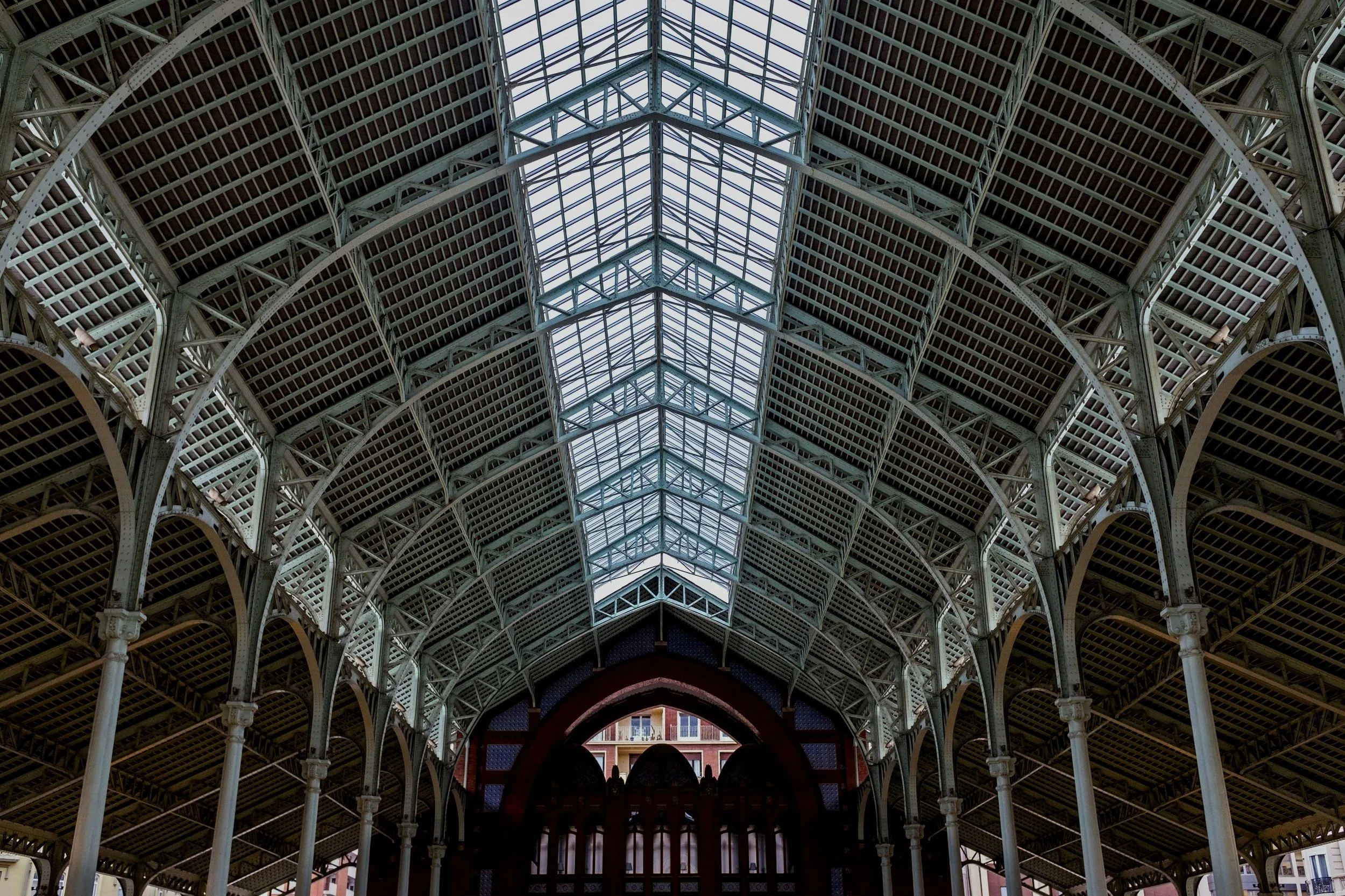 Interior view of a large metal and glass roof structure with arches and a central skylight, supported by white columns, with a building visible through an arch at the end.