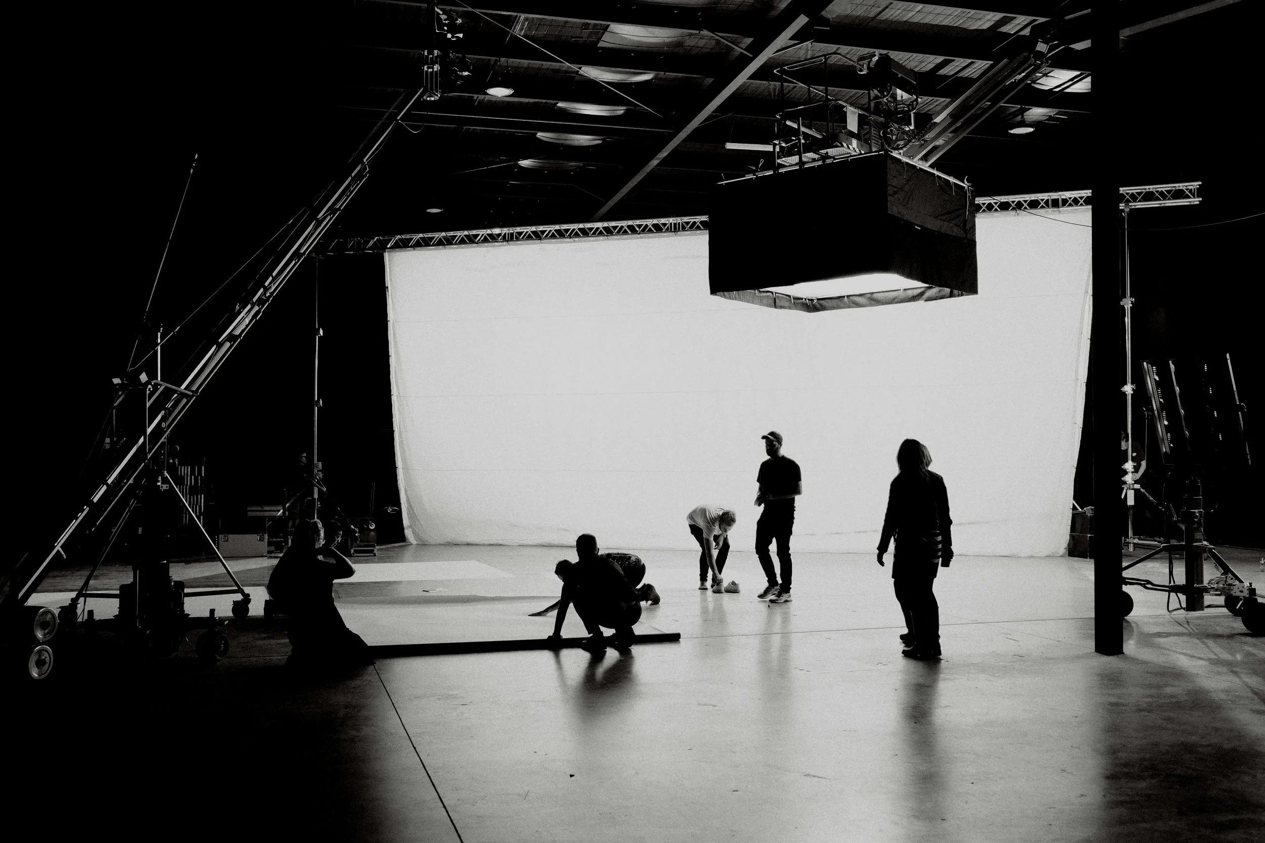 A film or photography studio with a large white backdrop, crew members setting up or adjusting equipment, and overhead lighting and rigging.