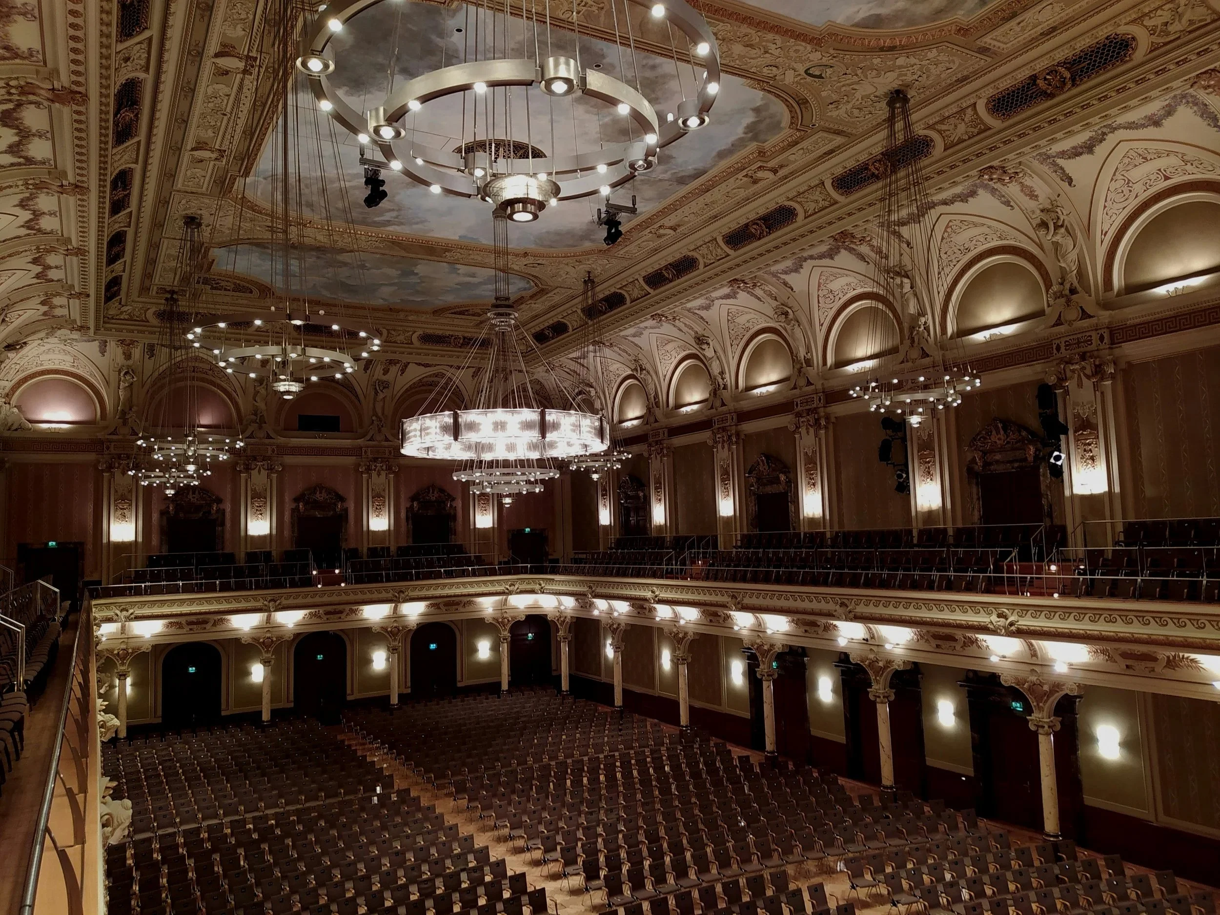 Interior of a grand, ornate theatre or concert hall with decorative arches, chandeliers, and rows of seats.