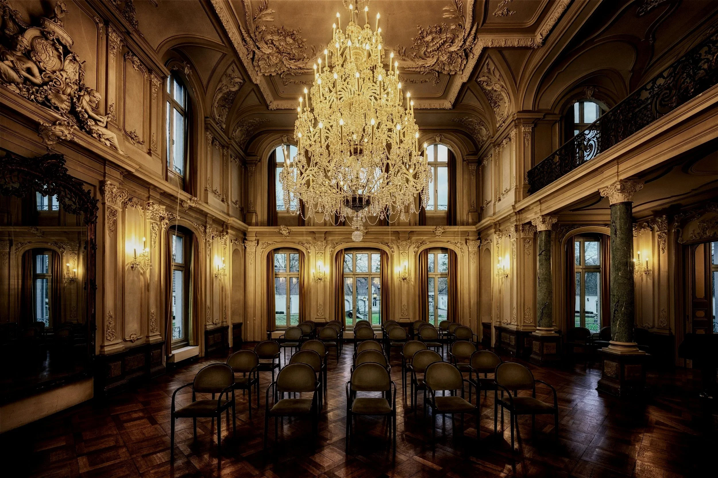 An elegant, ornately decorated room with a grand chandelier hanging from a high ceiling, tall windows with drapes, and multiple rows of chairs arranged for a meeting or event. The room features intricate molding, columns, and wood paneling.
