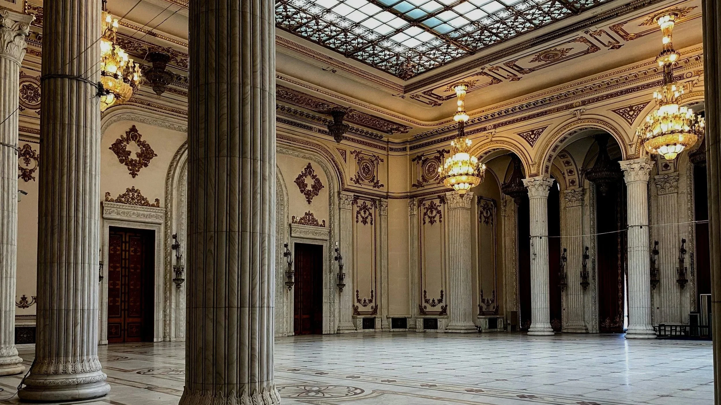 Interior of a grand hall with marble floors, large columns, ornate wall and ceiling decorations, and multiple chandeliers hanging from the ceiling.