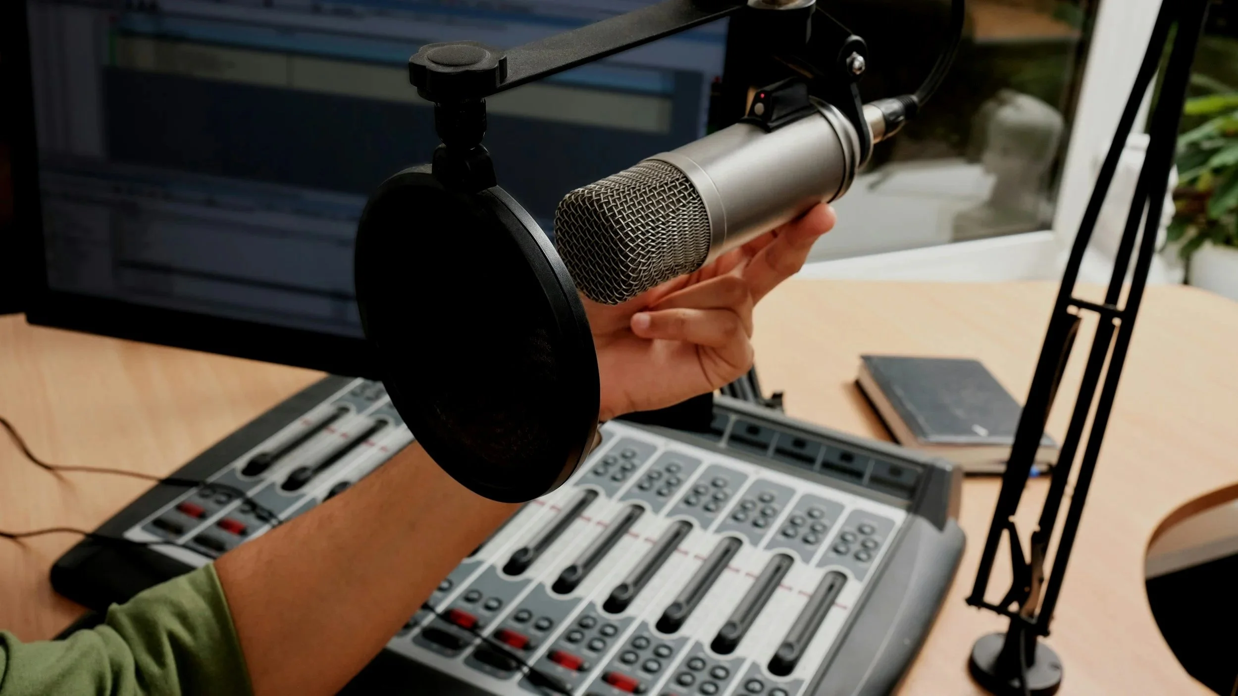 A hand holding a professional microphone with a pop filter in a radio studio setting, with audio mixing controls on a desk and a computer monitor in the background.