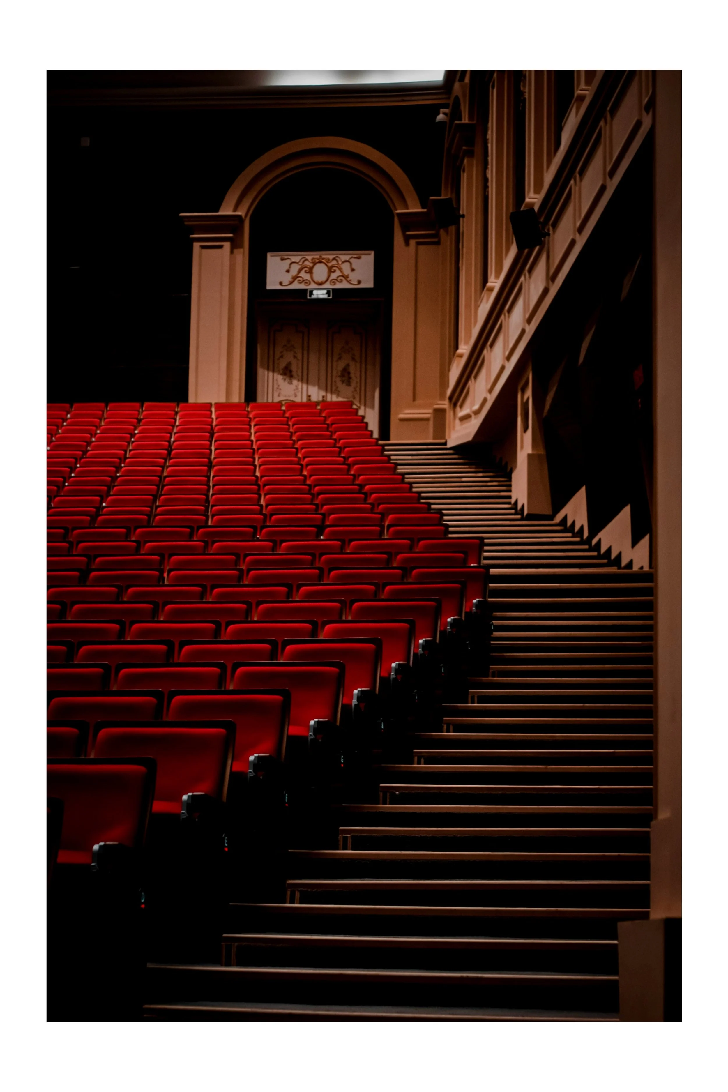 Empty theater with red seats, stairs, ornate decor, and a door at the top.