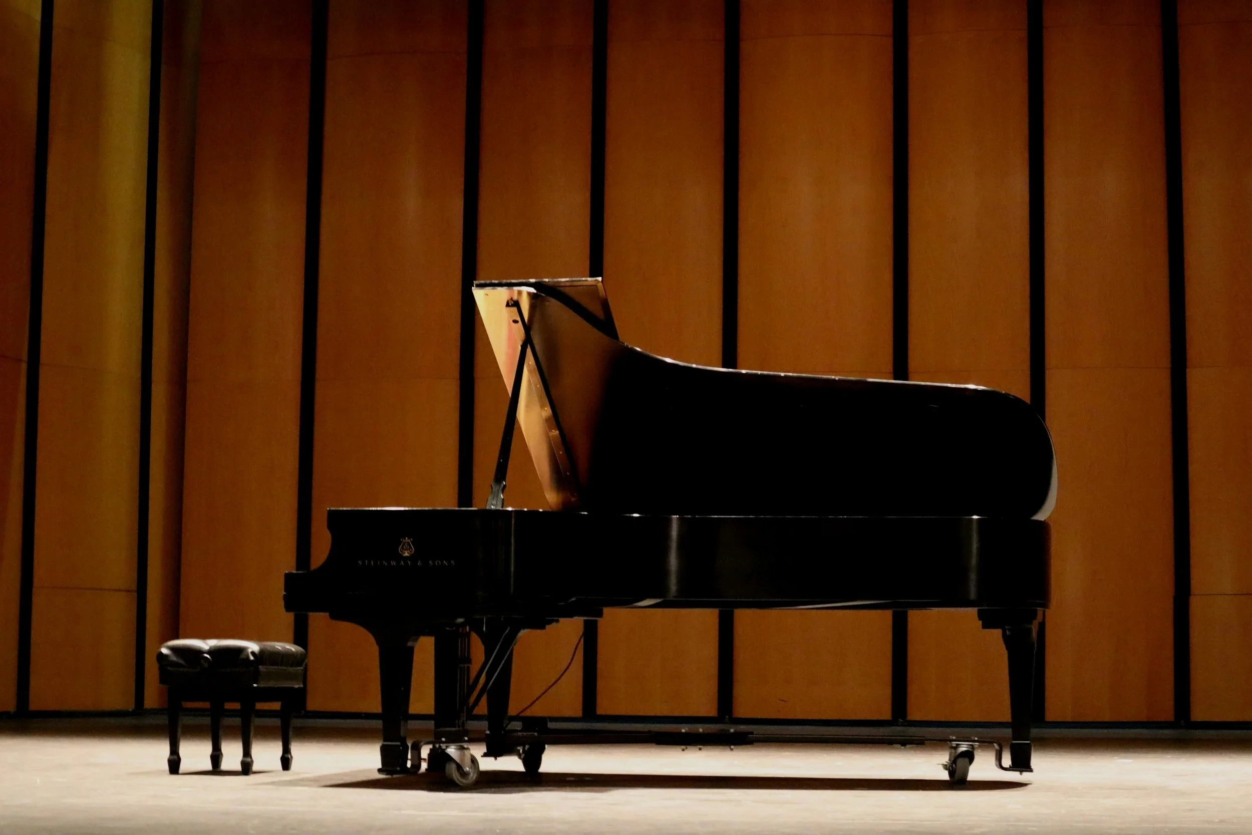 A grand black piano on a wooden stage with a matching piano bench, set against a wooden paneled wall.