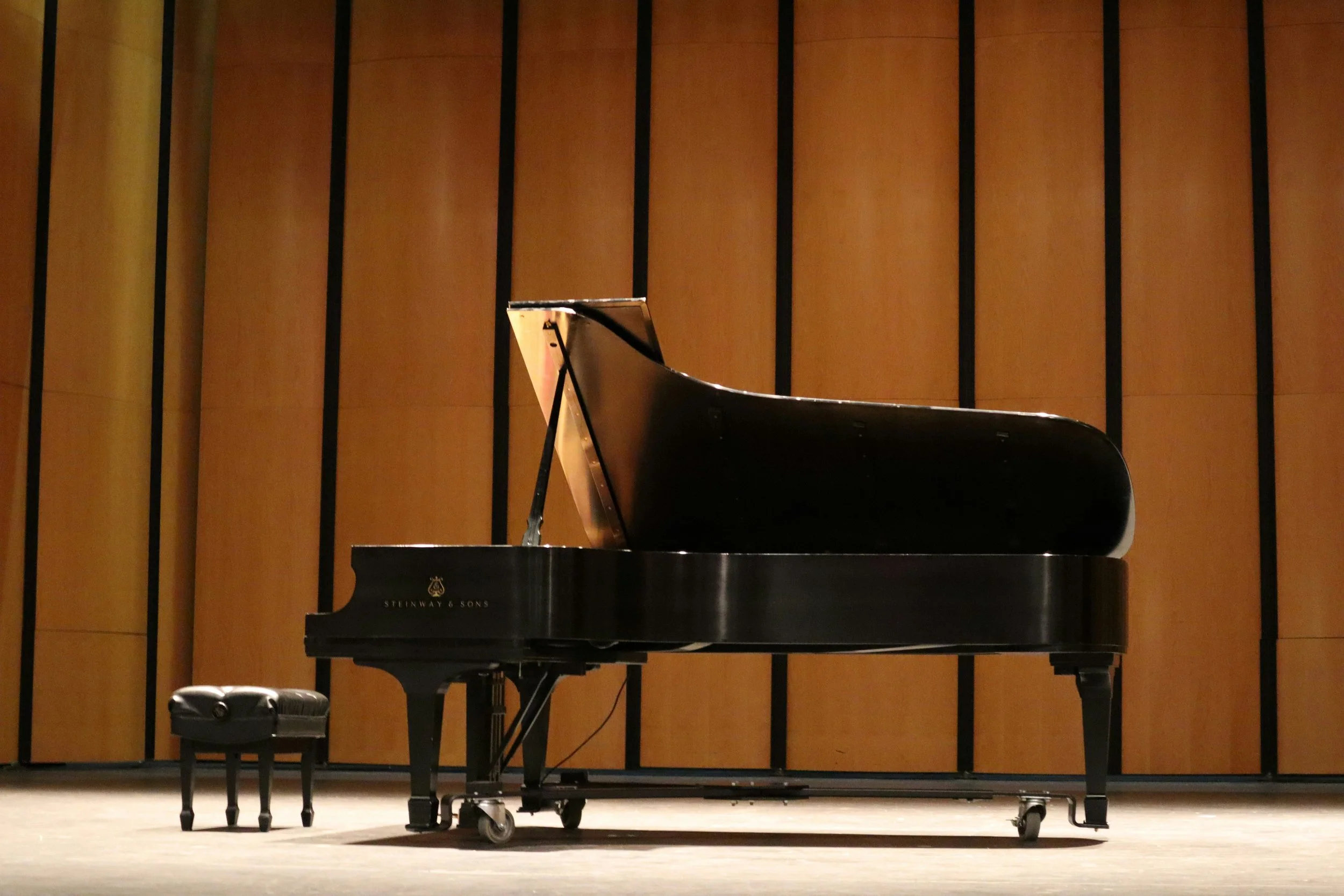 A black Steinway & Sons grand piano on a wooden stage with a matching bench, against a wooden wall with vertical black lines.