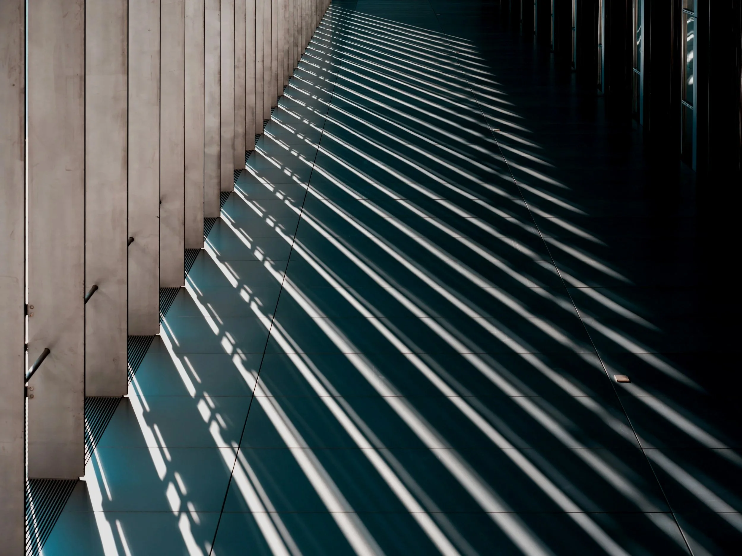 Light and shadow patterns of sunlight coming through slats cast on a modern building's exterior wall and floor.