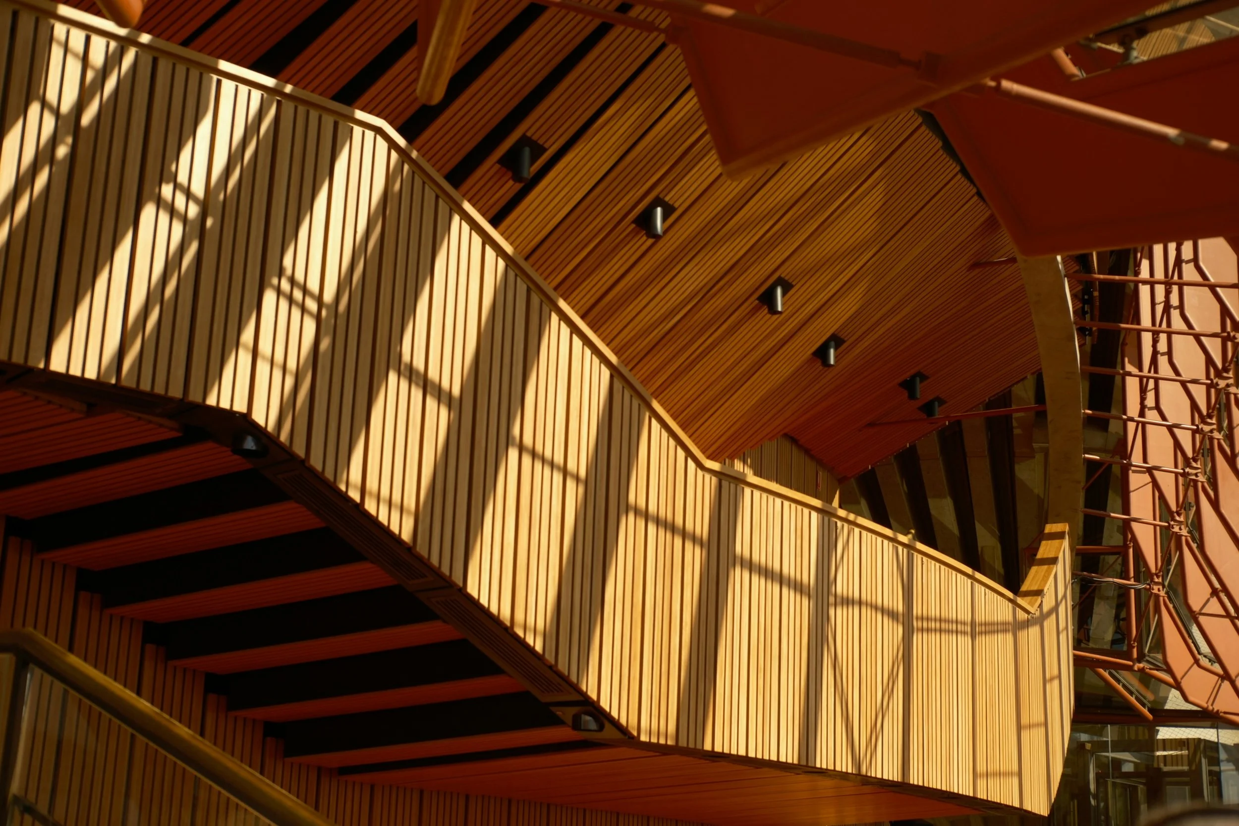 Interior view of a modern building with wooden stairs and railings, illuminated by sunlight and black ceiling lights.