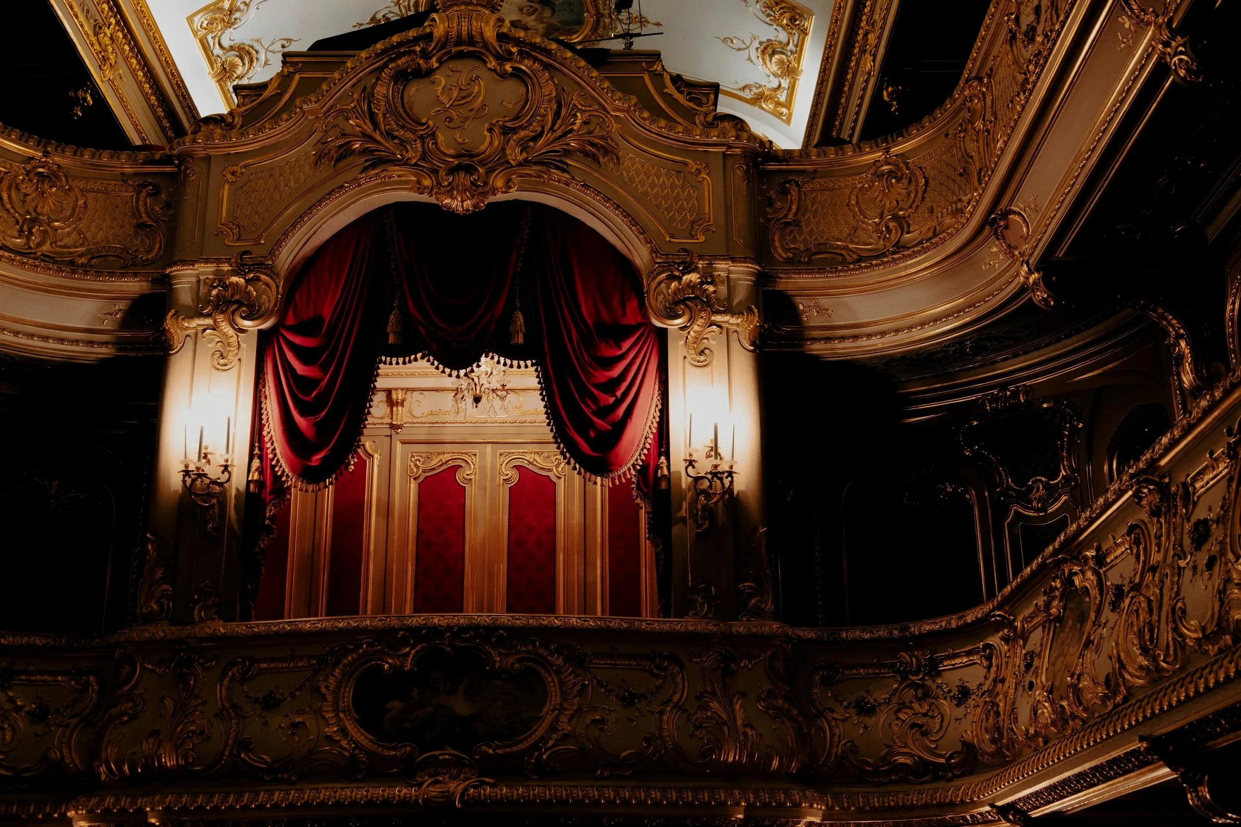 The ornate interior of an opera house or theater balcony with rich gold detailing, red velvet curtains, and a chandelier.