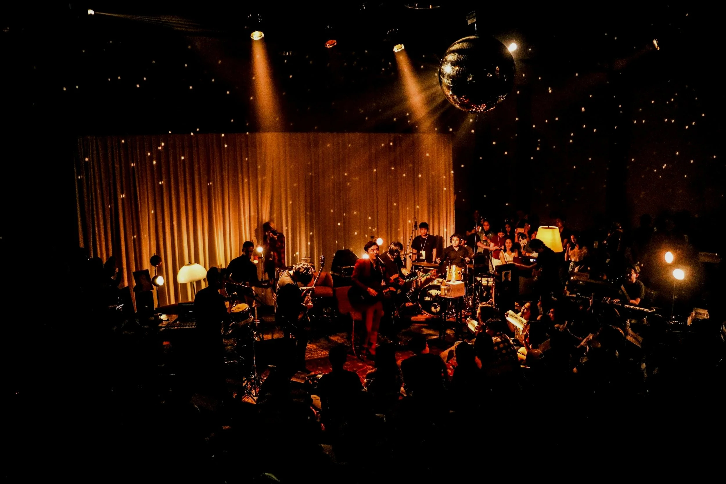 A live music performance on a dimly lit stage with a band playing instruments, a large golden curtain backdrop, warm lighting, a disco ball hanging from the ceiling, and an audience seated in front.