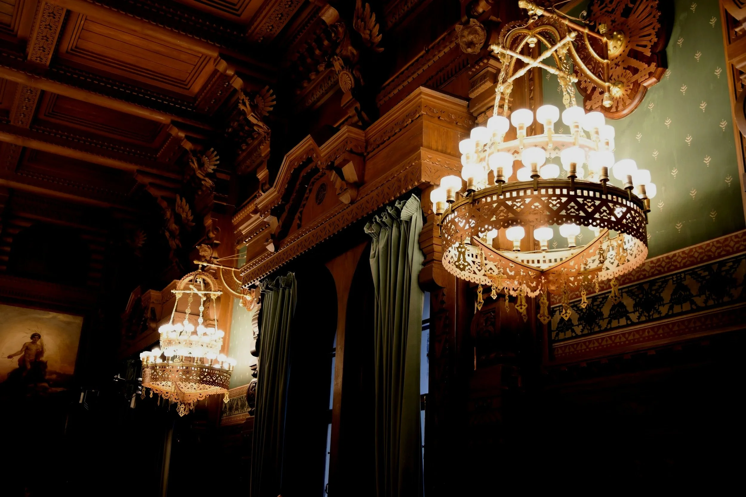 Intricate wood carvings and two elegant chandeliers illuminating a richly decorated ceiling and draped green curtains in a historic or luxurious interior.