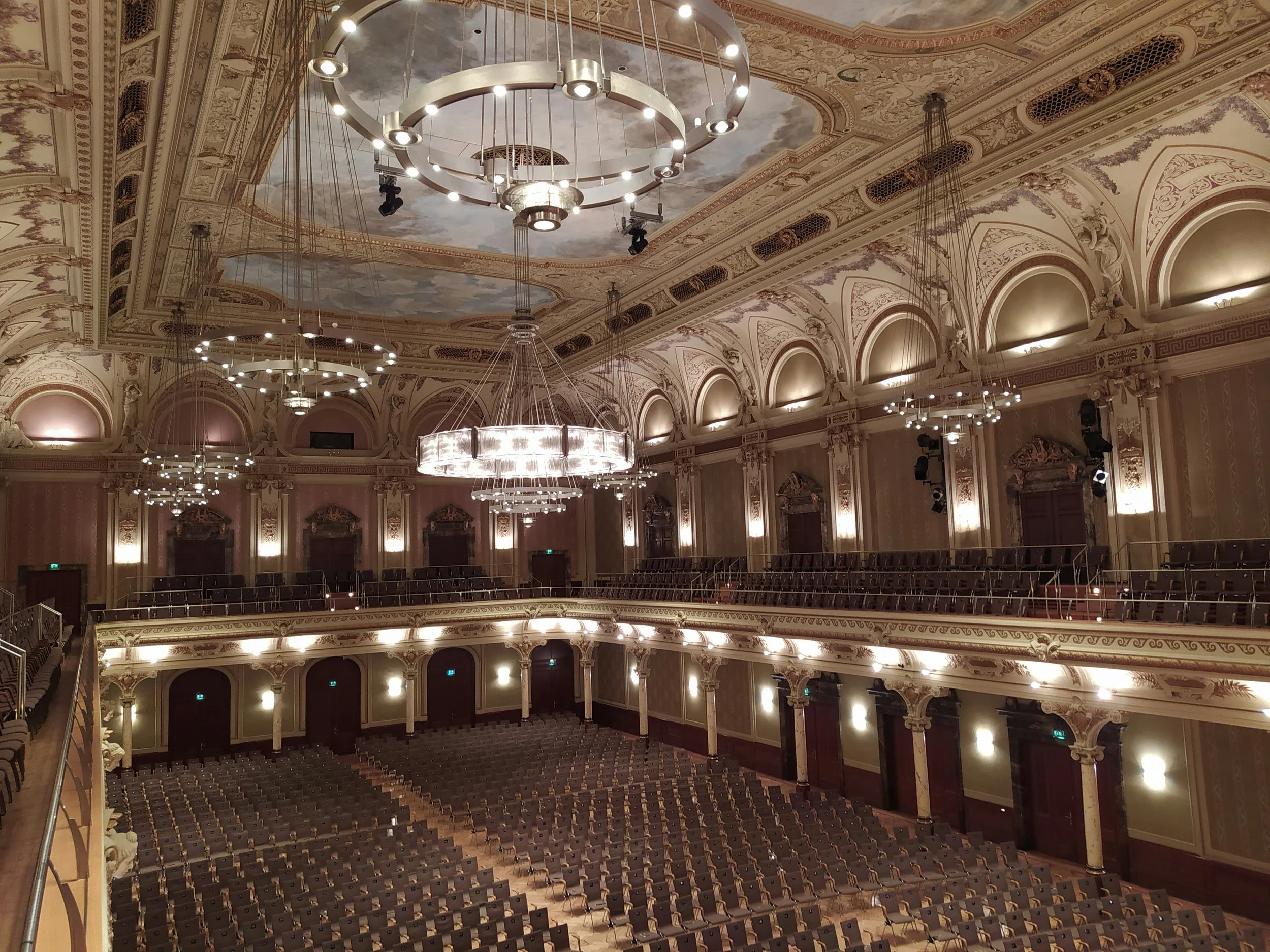 Empty ornate theatre hall with multiple chandelier lights, decorative ceiling, beige walls, and rows of folded chairs.