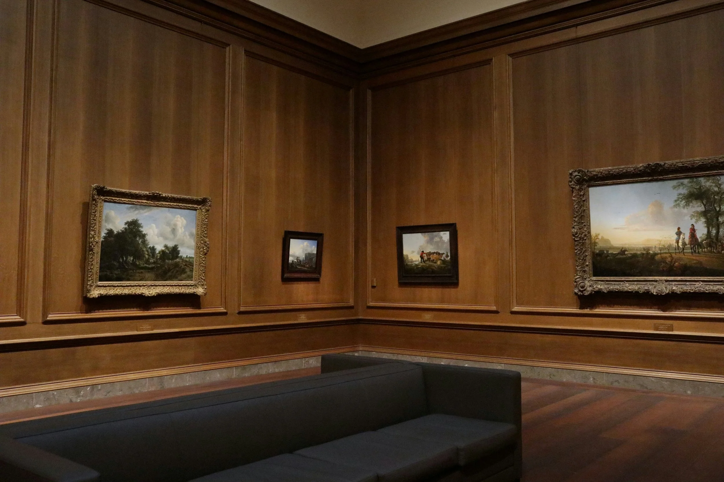 An art gallery with wooden-paneled walls, featuring four landscape paintings in ornate frames. A black couch is in the foreground.