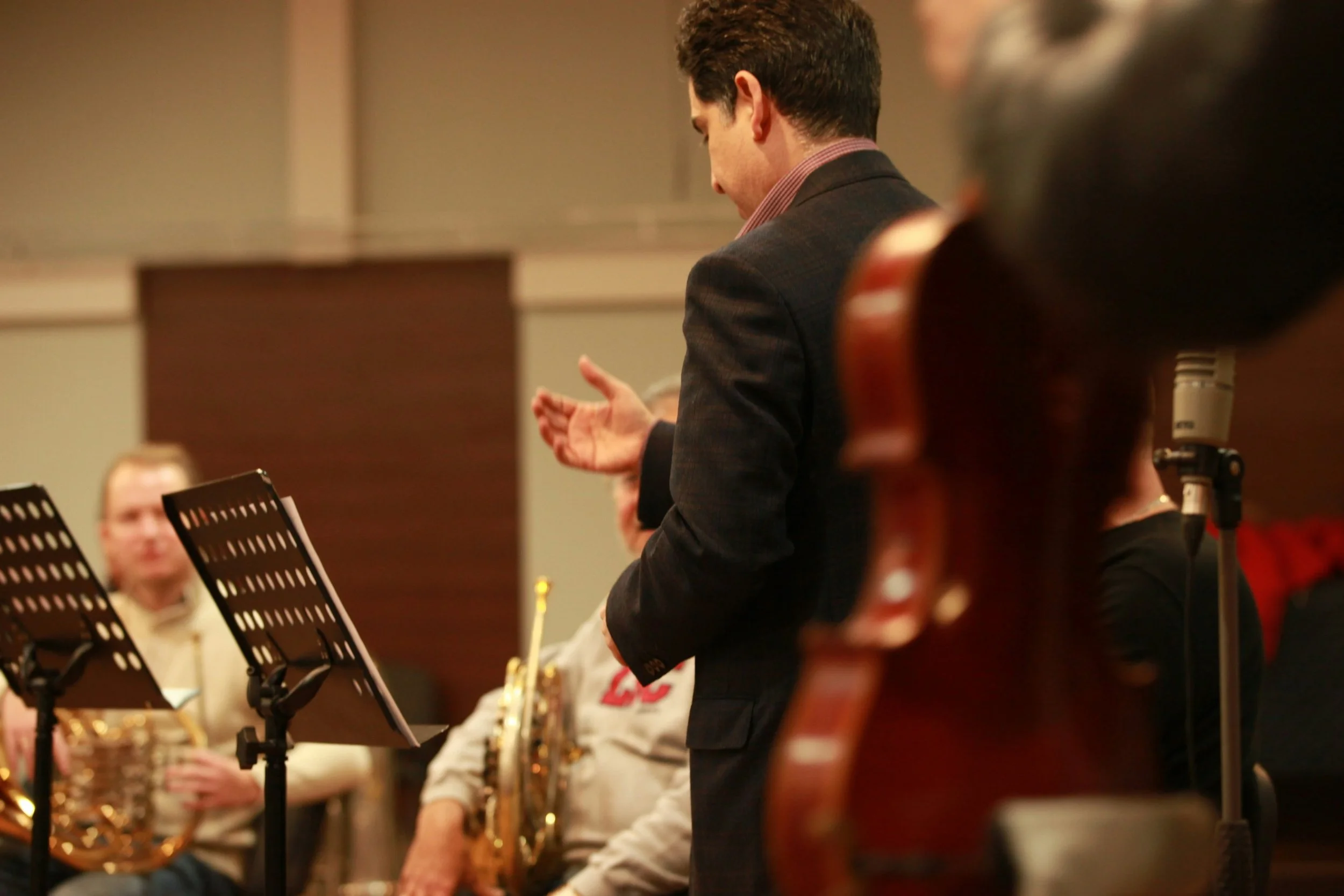 A man in a dark blazer conducting an orchestra, with musicians in the background holding brass instruments, in a concert hall.