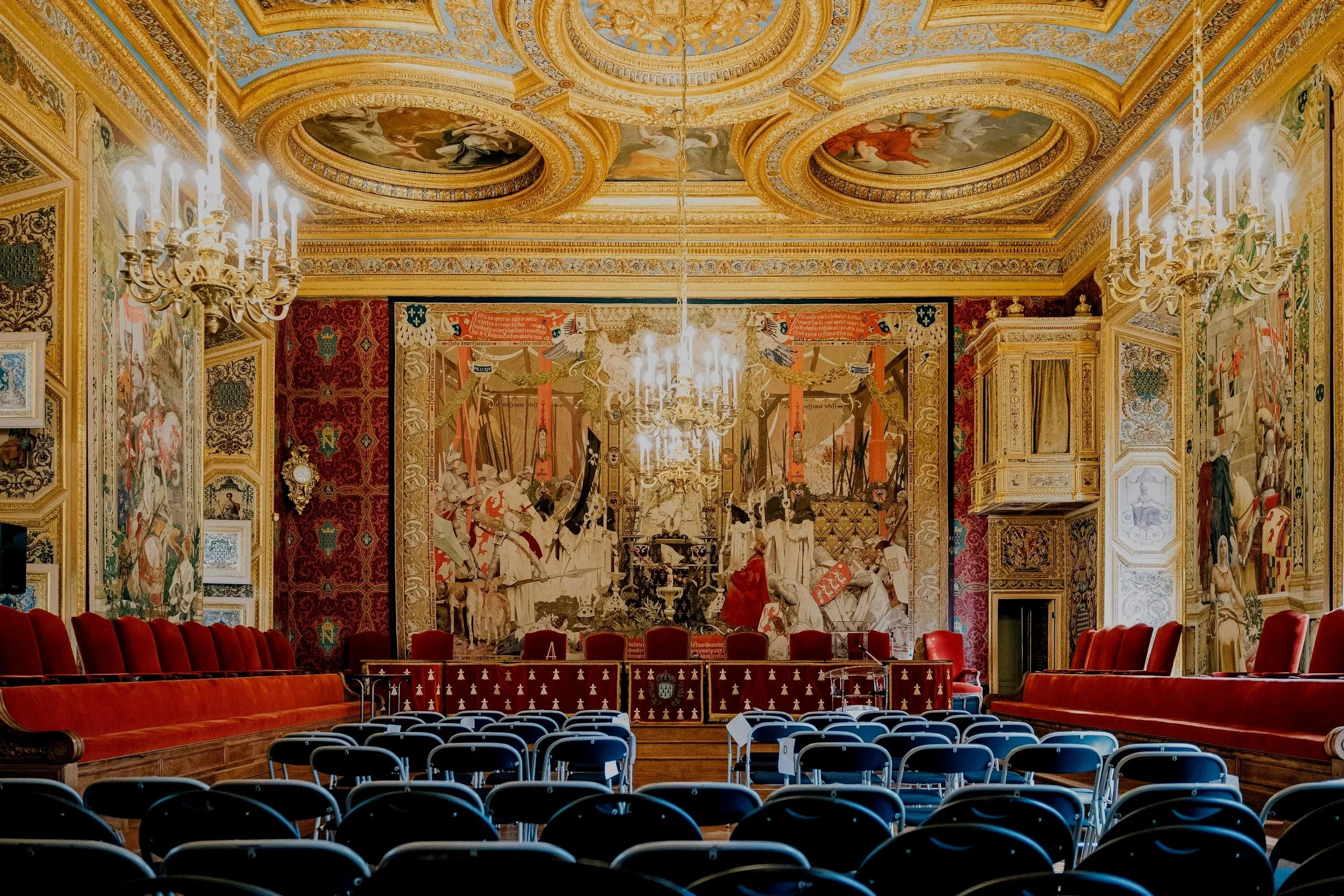 A grand ornate room with gold and red decor, chandeliers, and a mural on the wall. The room has rows of chairs facing a long table with red velvet chairs.