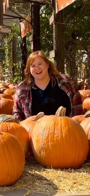 A young woman with red hair and a plaid shirt smiling among pumpkins at a pumpkin patch.