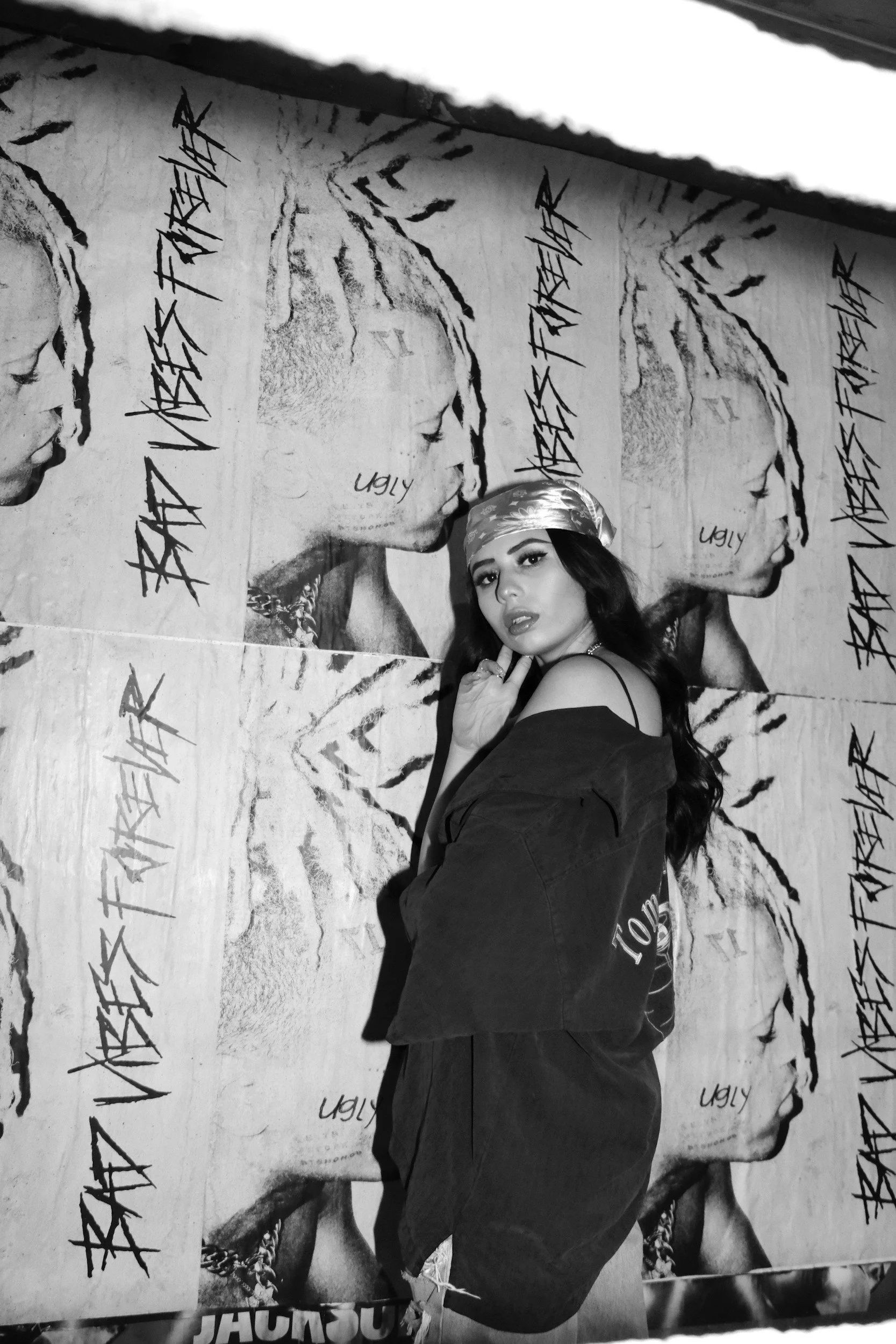 A woman with dark hair and a bandana poses in front of a repeating poster featuring a woman's profile, with the words 'Ugly' and stylized text.