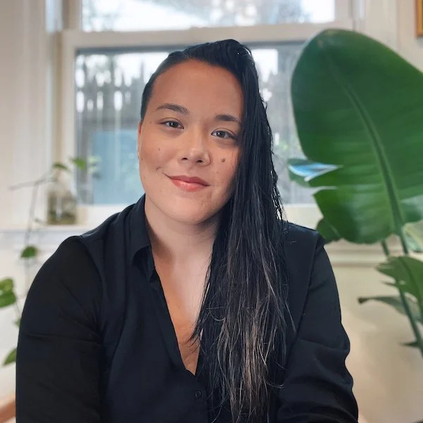 A non-binary AFAB person with long dark hair, wearing a black shirt, sitting in front of a window with green plants around her.