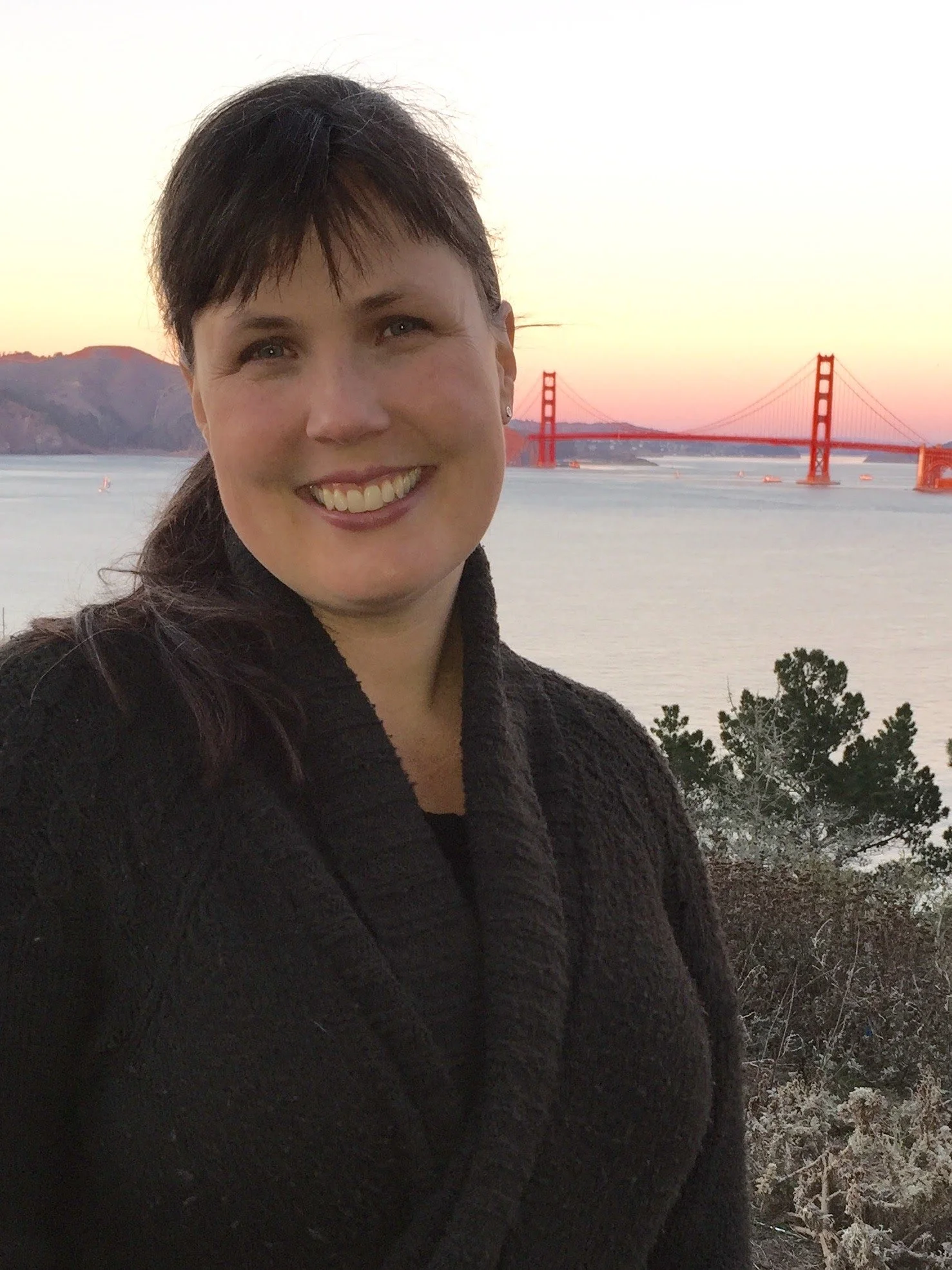 A woman smiling outdoors with the Golden Gate Bridge in the background during sunset.