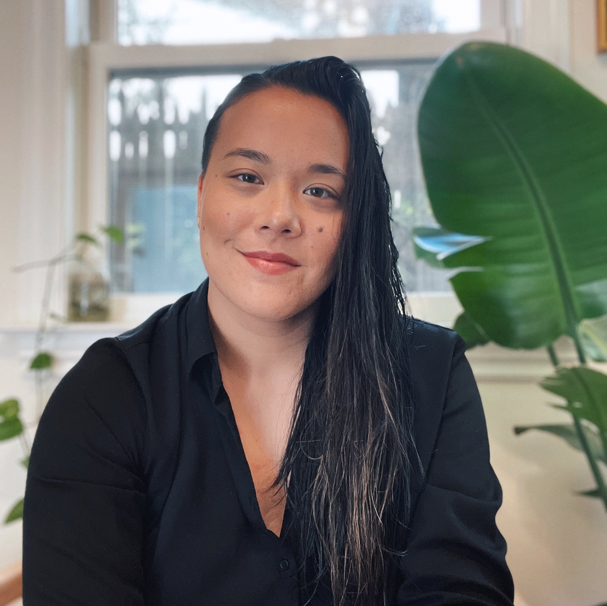 A non-binary AFAB person with long black hair, wearing a black top, sitting indoors near a window with sunlight streaming in, partially obscured by large green plant leaves in the foreground.