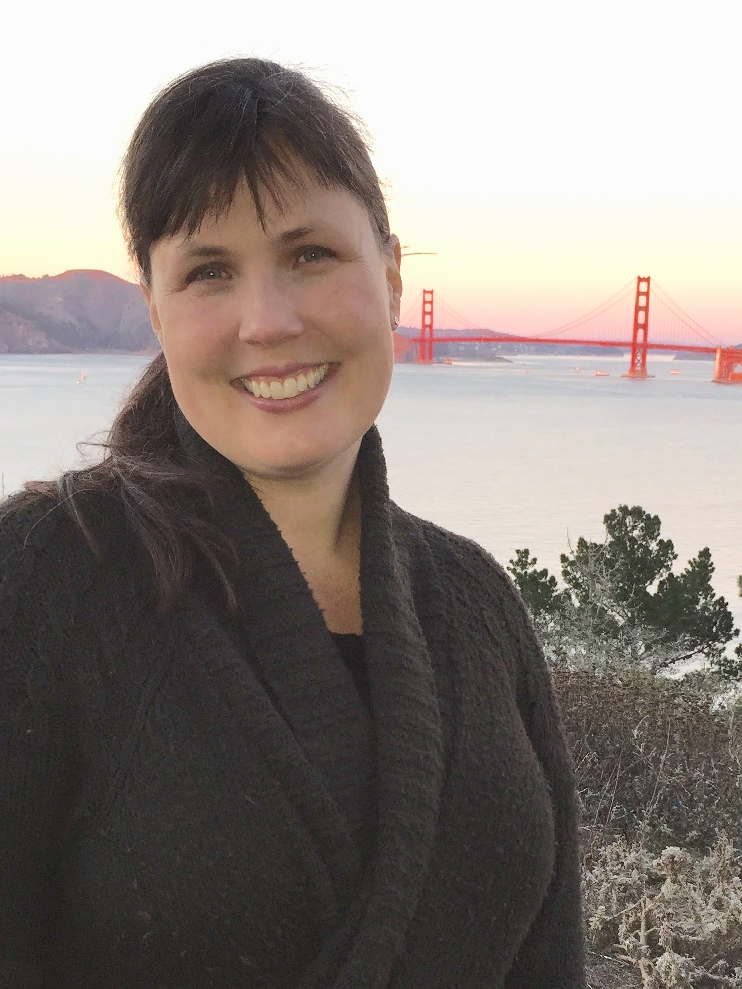 A woman smiling in front of the Golden Gate Bridge during sunset, with hills and trees in the background.