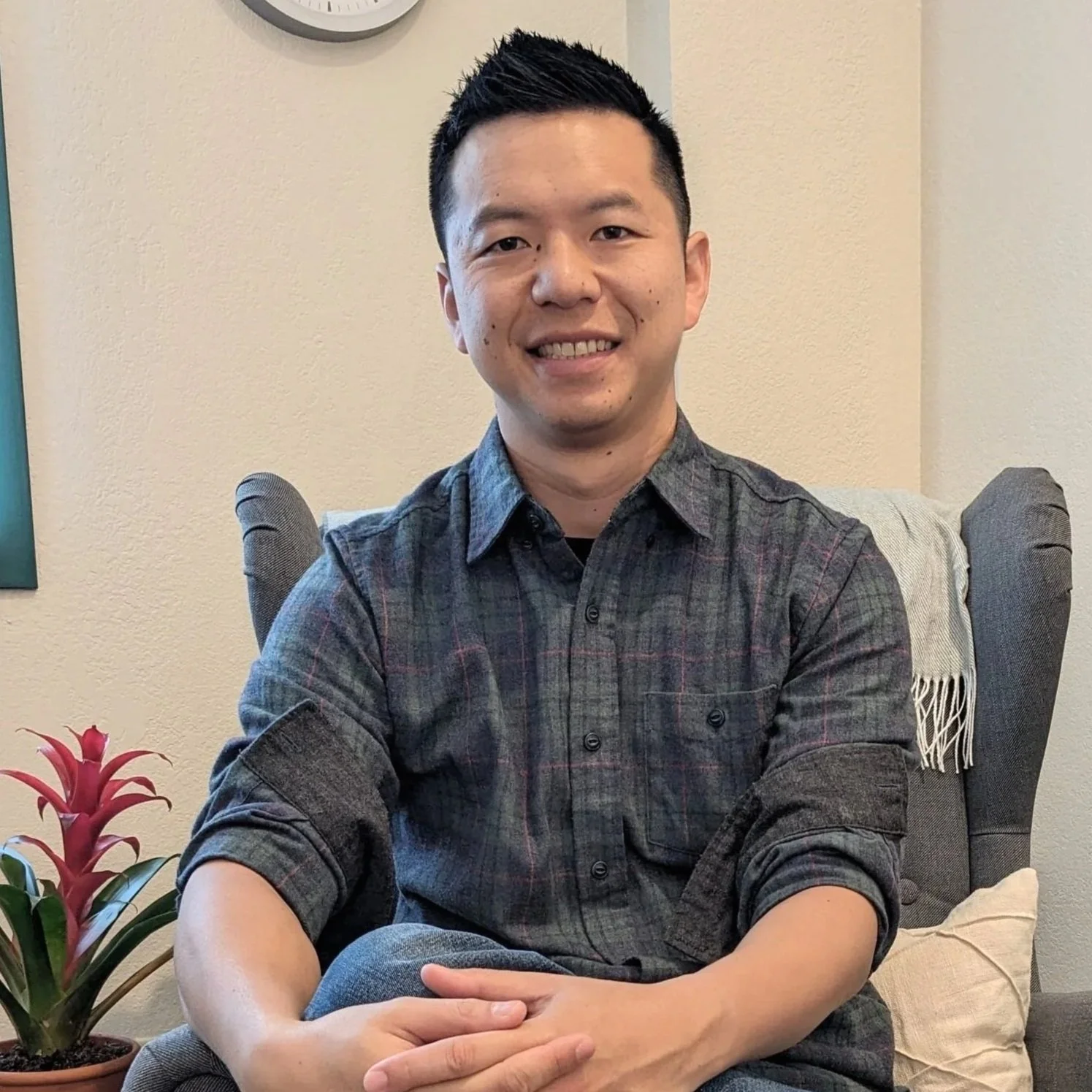 A smiling man with short dark hair and a plaid dark shirt sitting on a gray armchair with a cream pillow behind him, a beige wall, a clock, and a plant with pink flowers in the foreground.