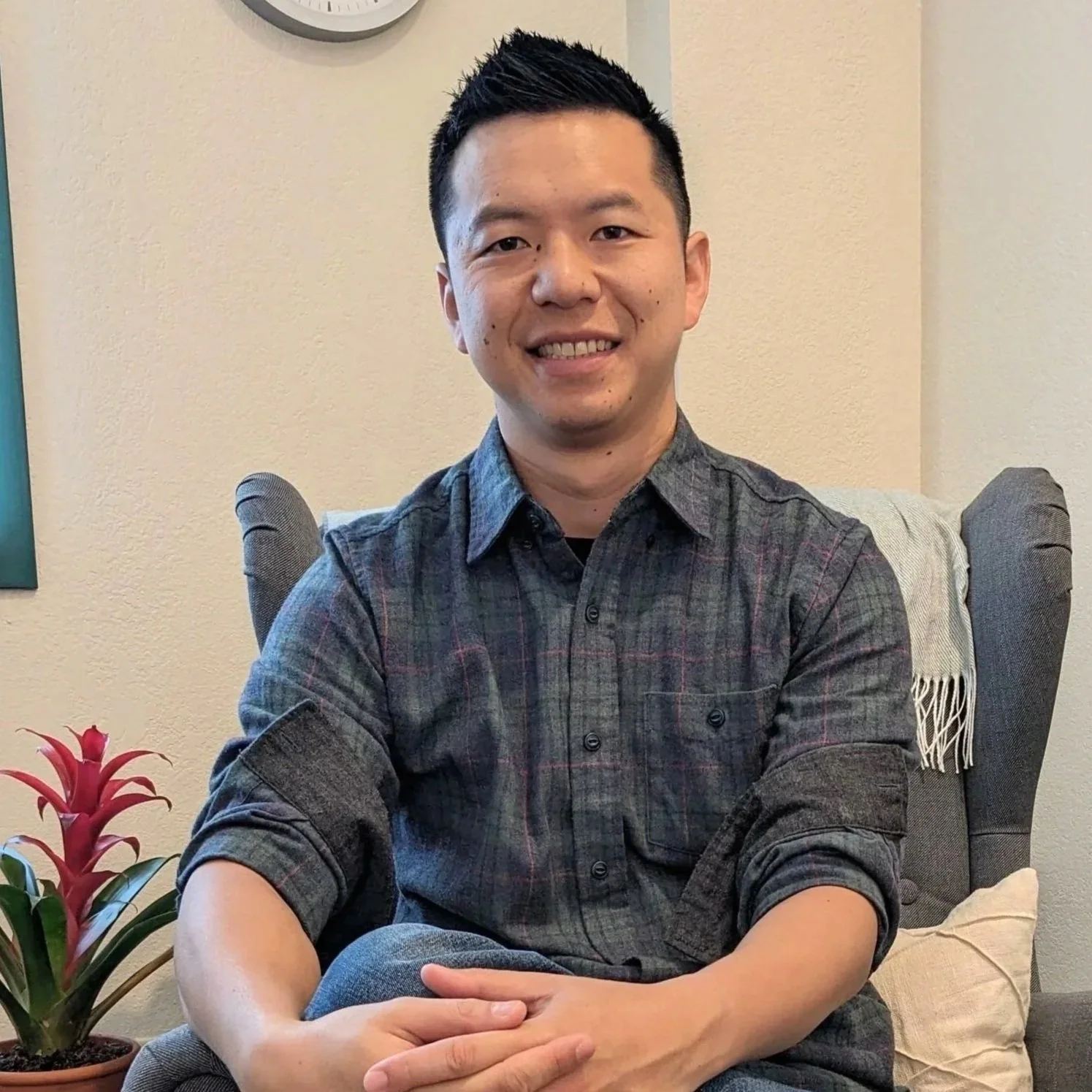 A man with black hair and a cheery smile, wearing a gray plaid shirt, sitting on a comfortable chair with a cream-colored pillow, beside a red plant, in a cozy indoor setting.
