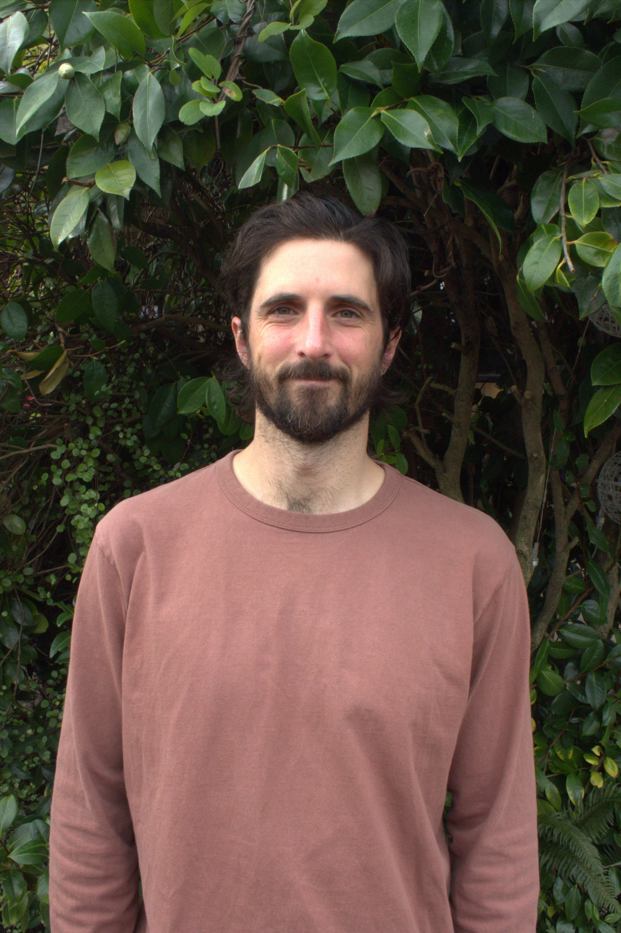 A man with dark hair and a beard, wearing a brown long-sleeve shirt, standing in front of a leafy green background.