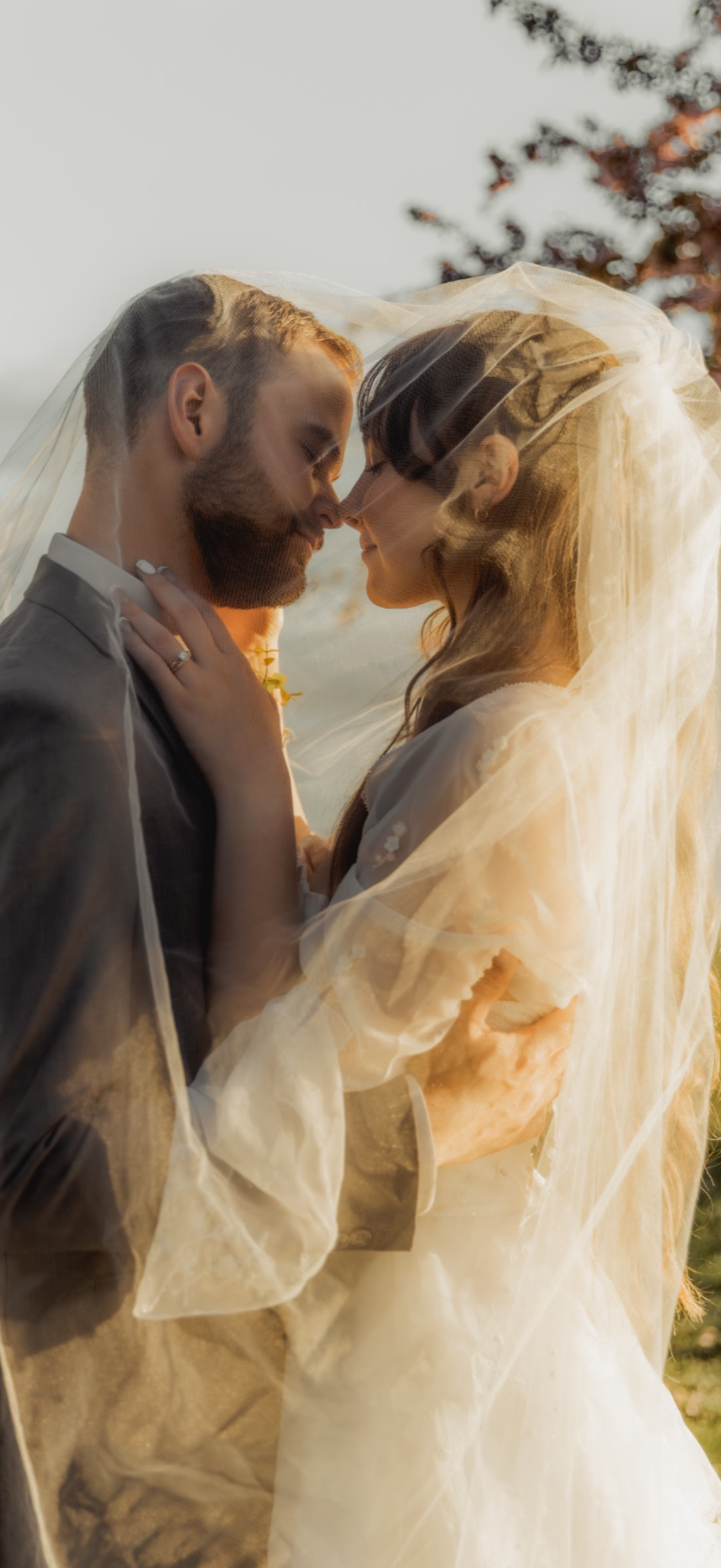 A bride and groom with closed eyes, touching foreheads, wrapped in a veil during their wedding outdoors at sunset. Montana Wedding Den Bosch Events. Photo by Cailee Kelly photography