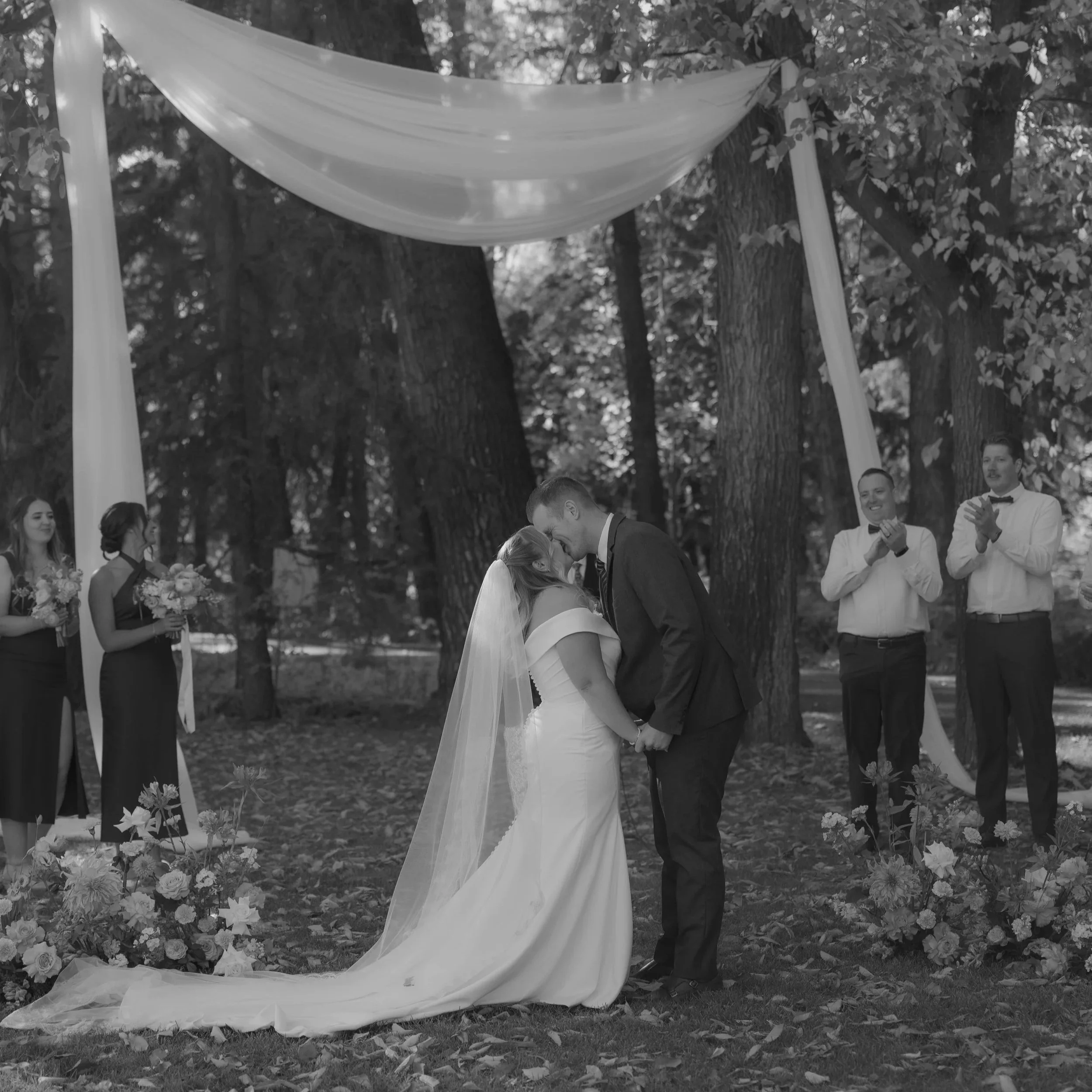 Black and white photo of a wedding ceremony outdoors with a bride and groom kissing, surrounded by bridesmaids and groomsmen, and large trees in the background.