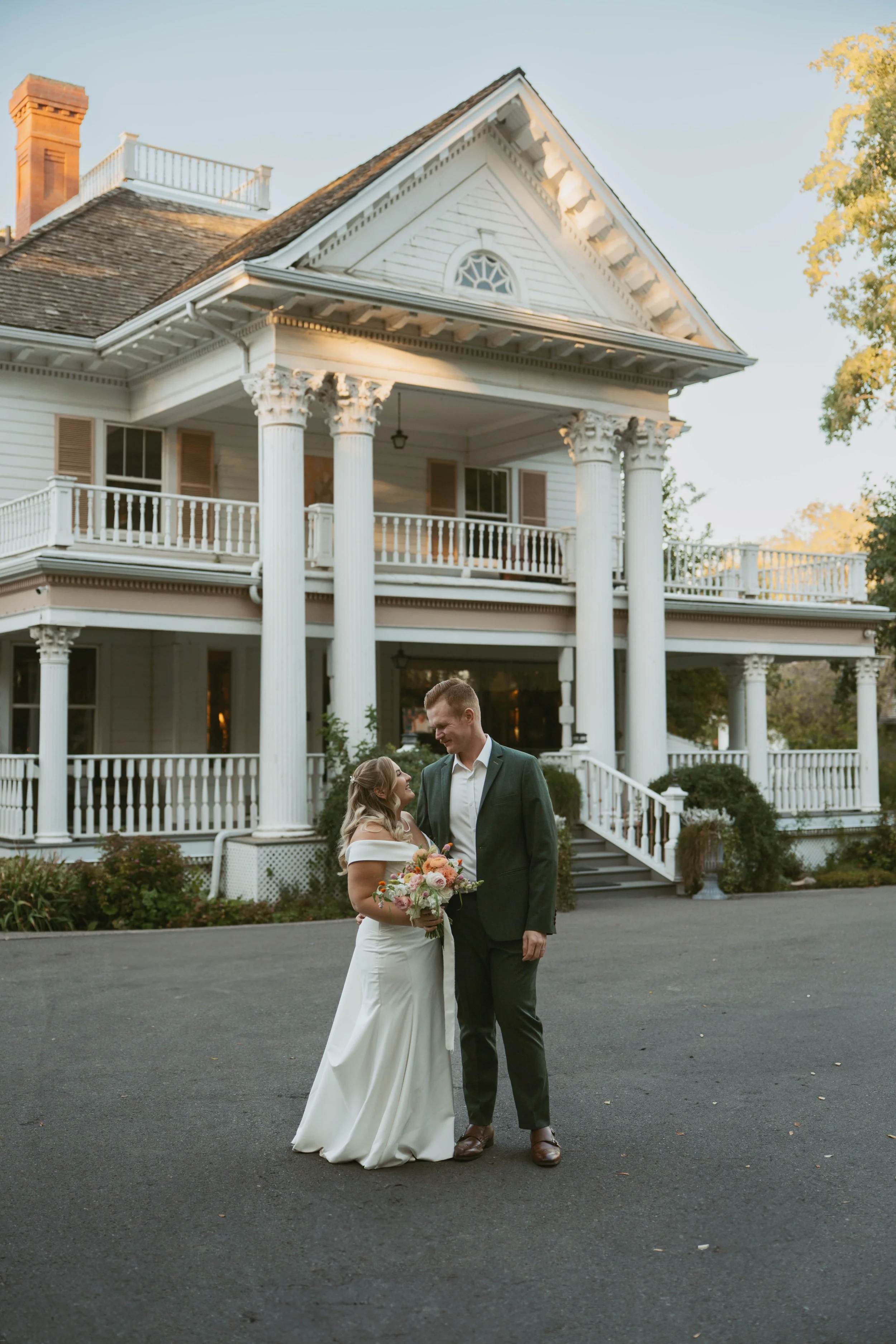 A newlywed couple stands on a paved driveway in front of Norland Historic Estate , Victorian-style house. Wedding Planned by Den Bosch Events and photographed by Juniper Bee Photography