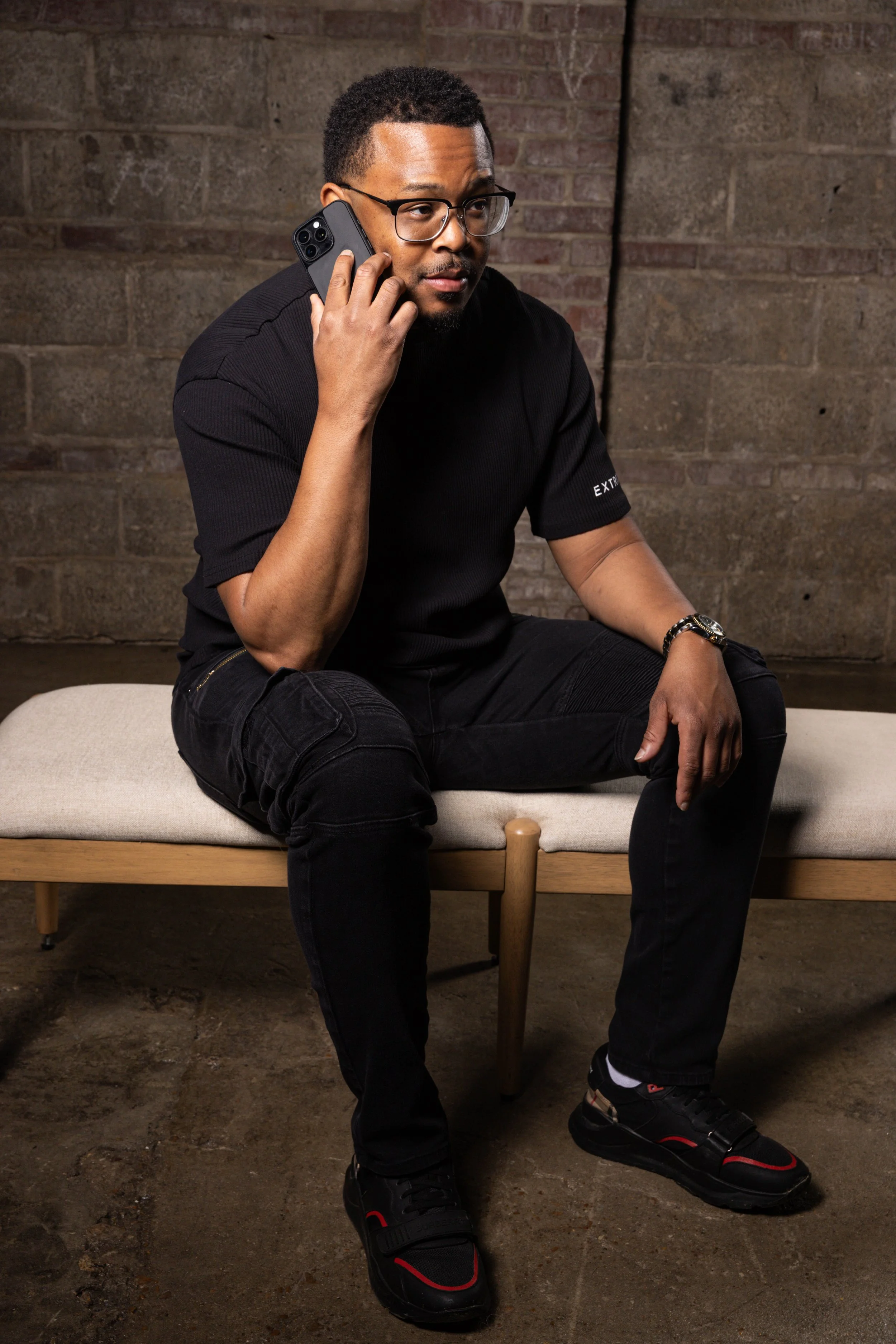 A man sitting on a bench against a brick wall, wearing glasses, a black T-shirt, black jeans, and black sneakers, while talking on his cell phone.