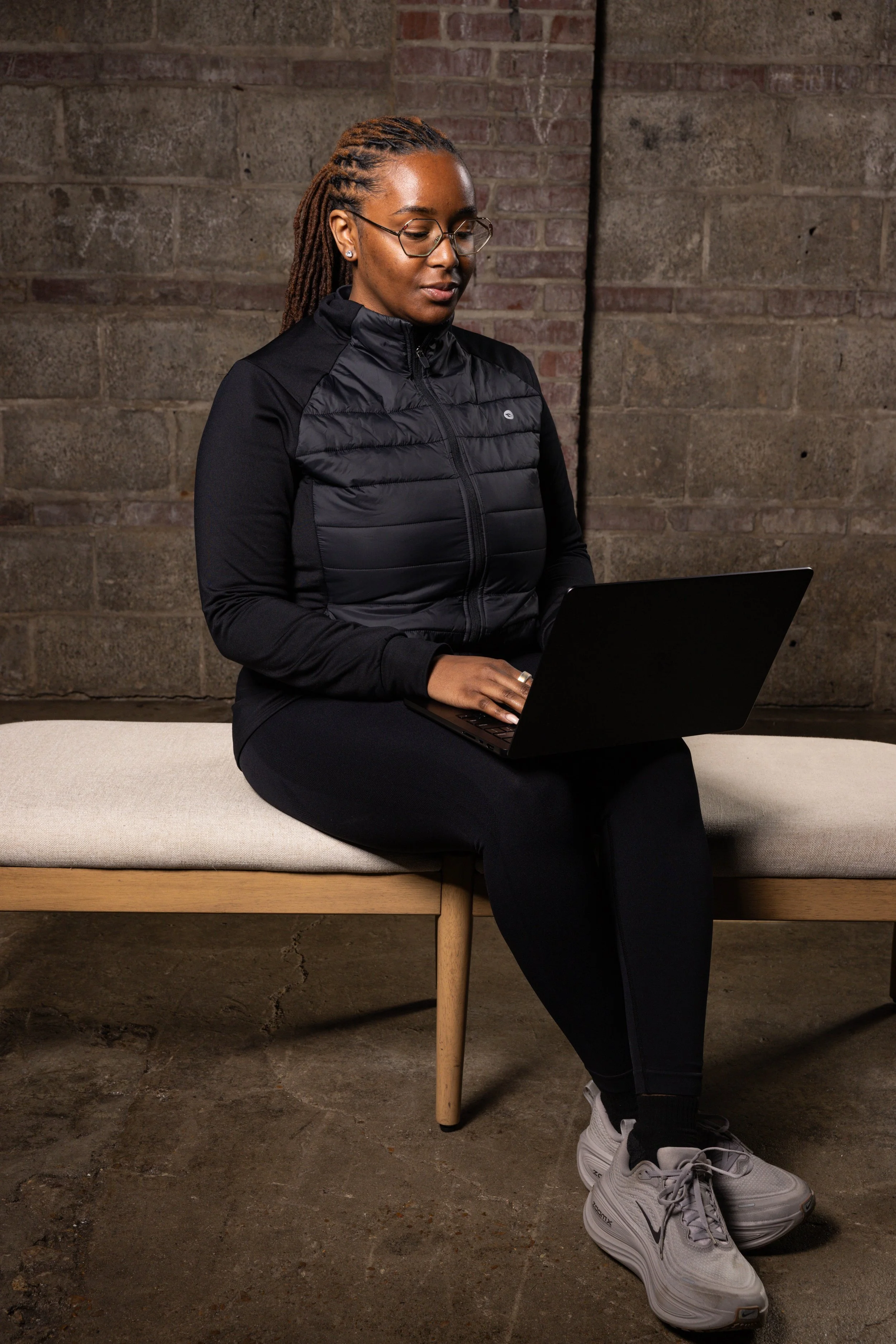 A woman with glasses and dreadlocks sitting on a beige bench, working on a laptop, against an exposed brick wall.