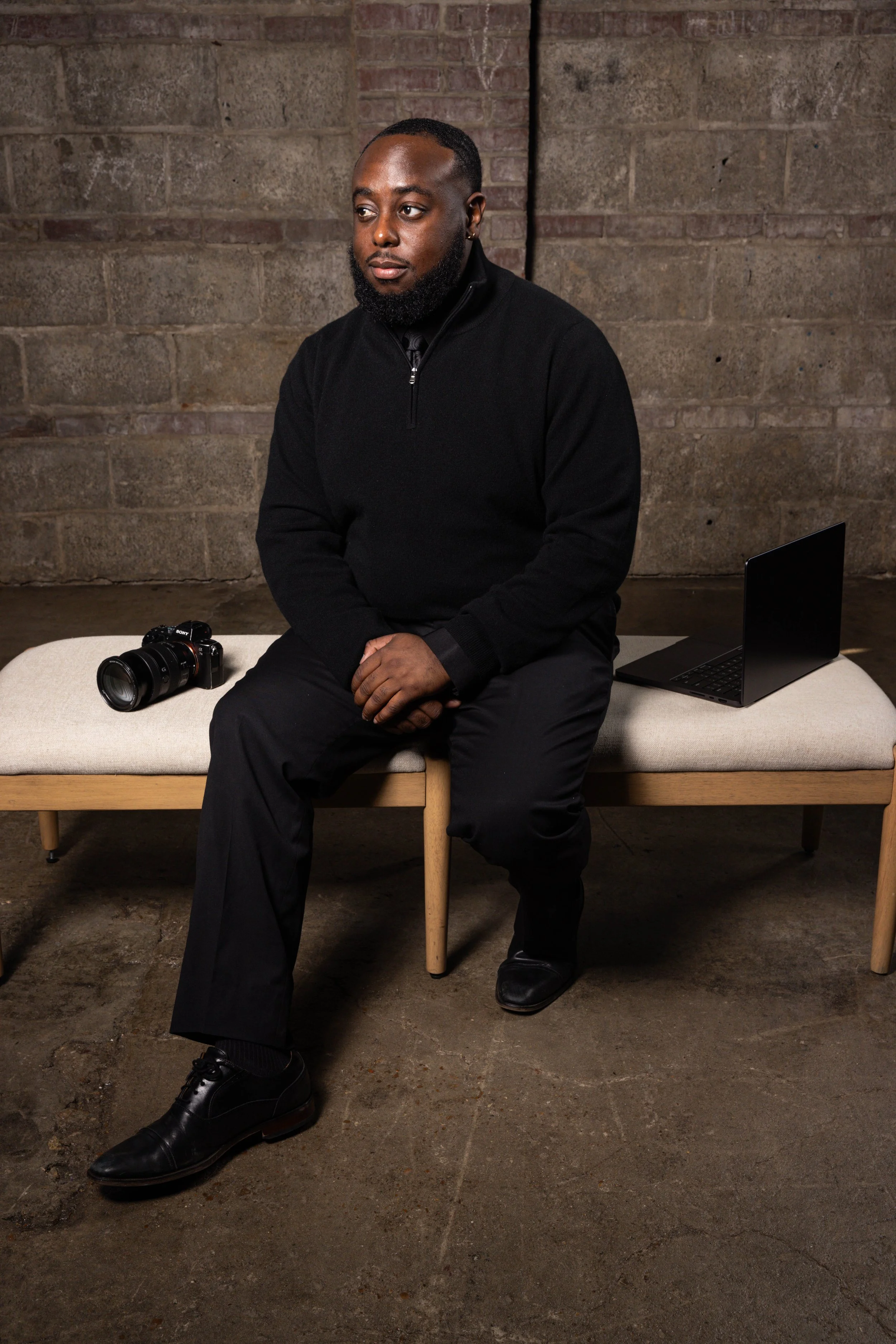 A man in black clothing sitting on a bench against an industrial brick wall background. There is a camera on the bench to his left and a laptop to his right.