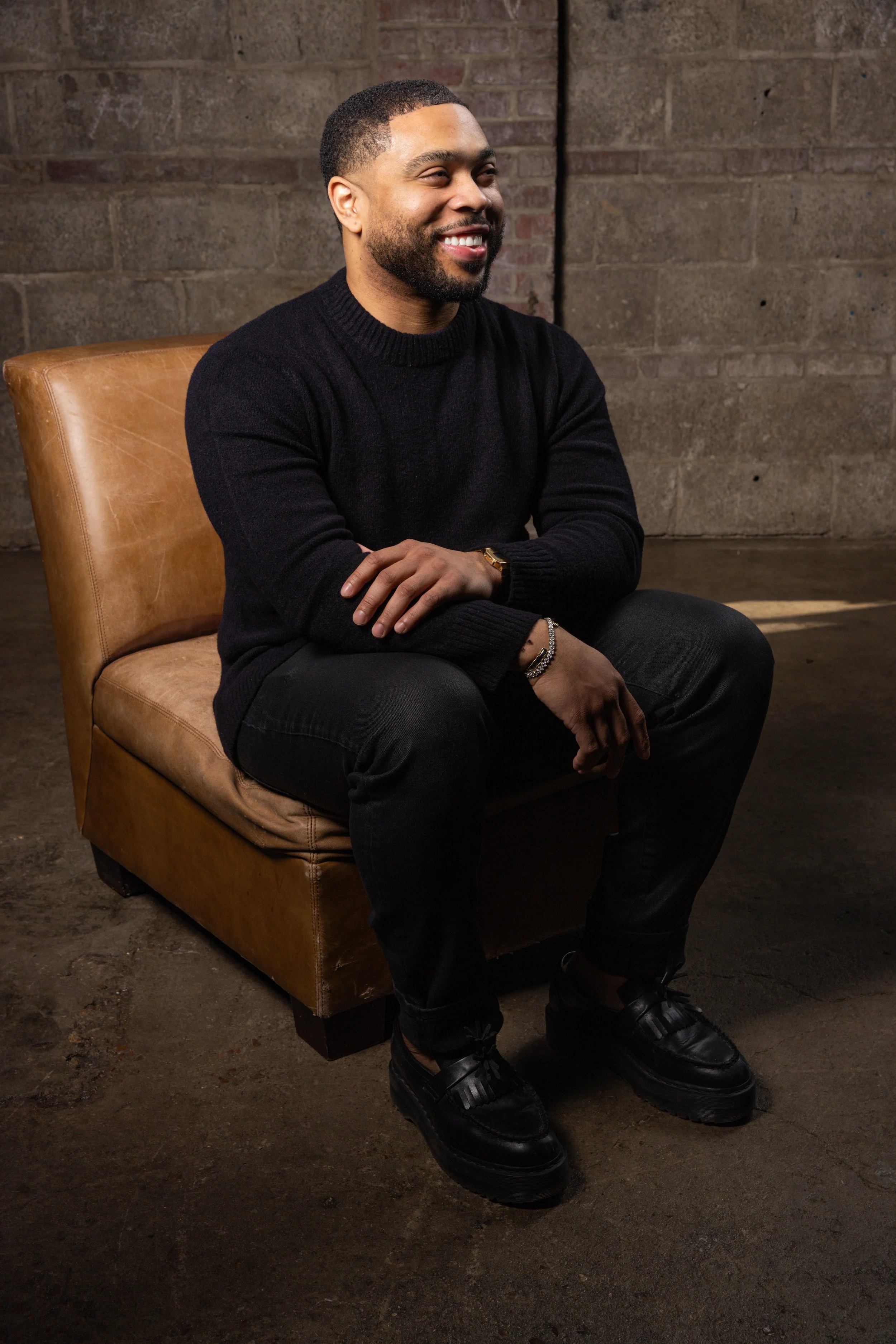 A man with a beard and short hair, wearing a black sweater, black pants, and black shoes, sitting on a tan leather chair against a brick wall, smiling and looking to his right.
