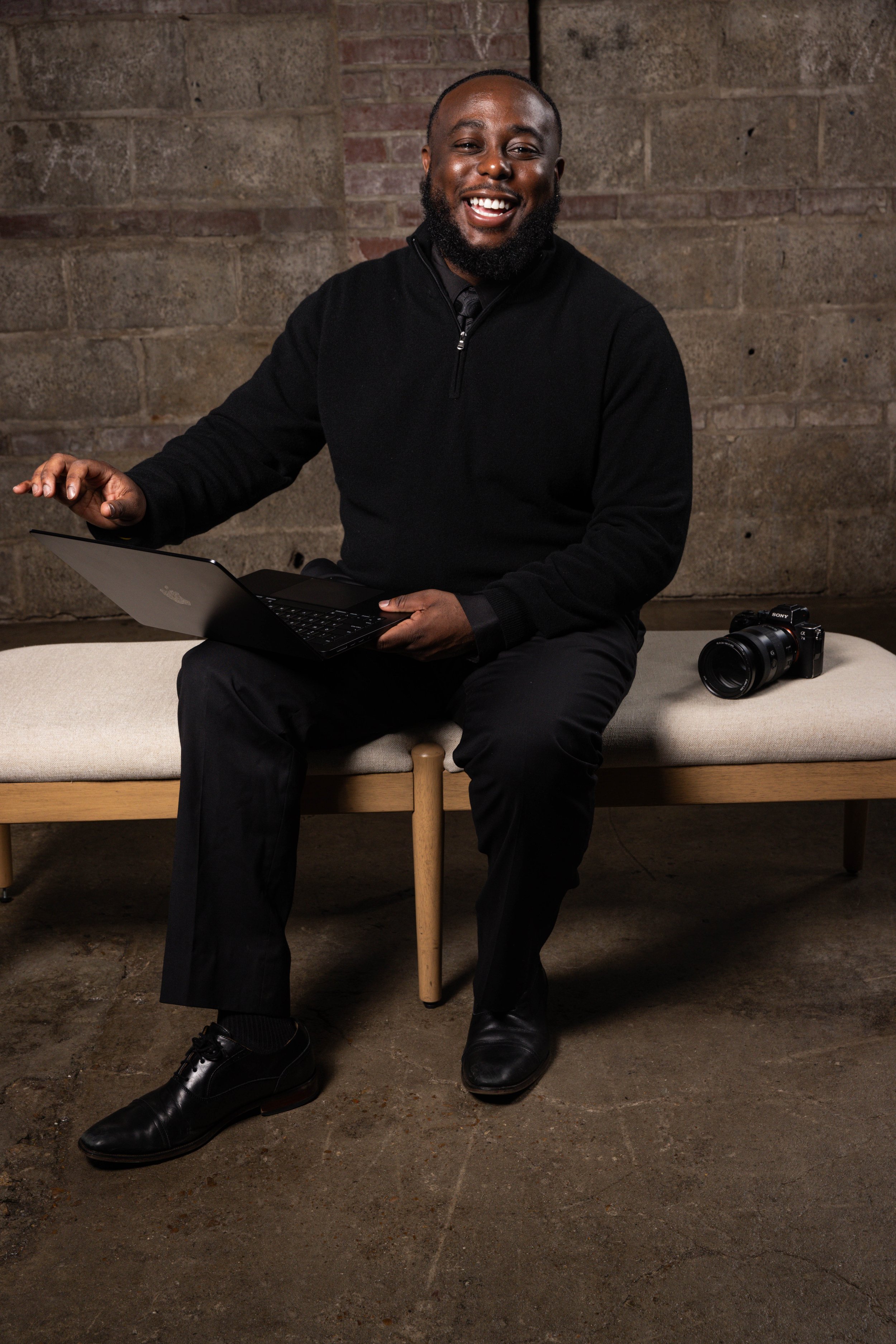 A smiling man with a beard sitting on a beige bench, using a laptop, with a camera placed beside him, against a brick wall background.